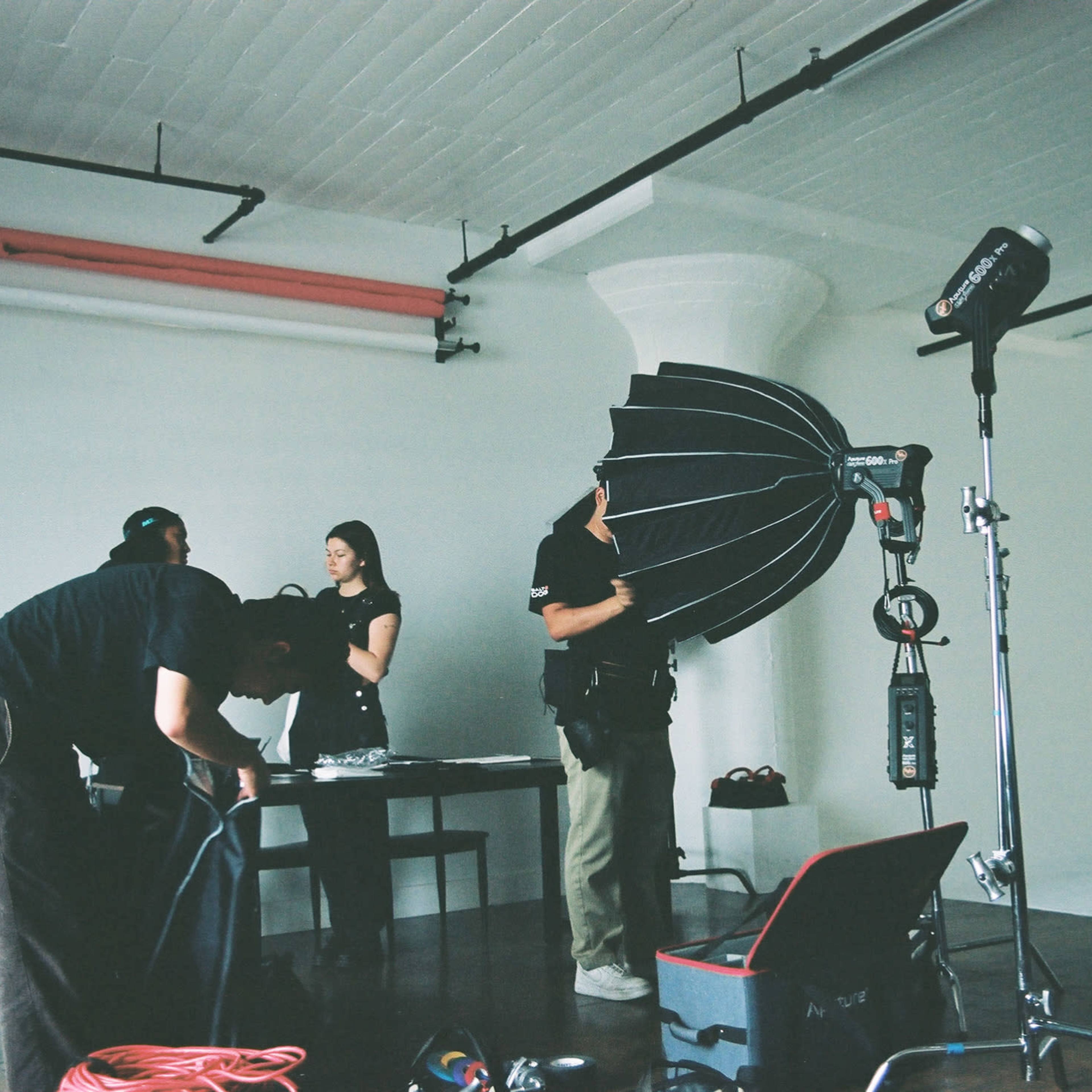 A group of people is setting up equipment in a minimalist studio space for a photoshoot, featuring a large umbrella softbox and a ladder.