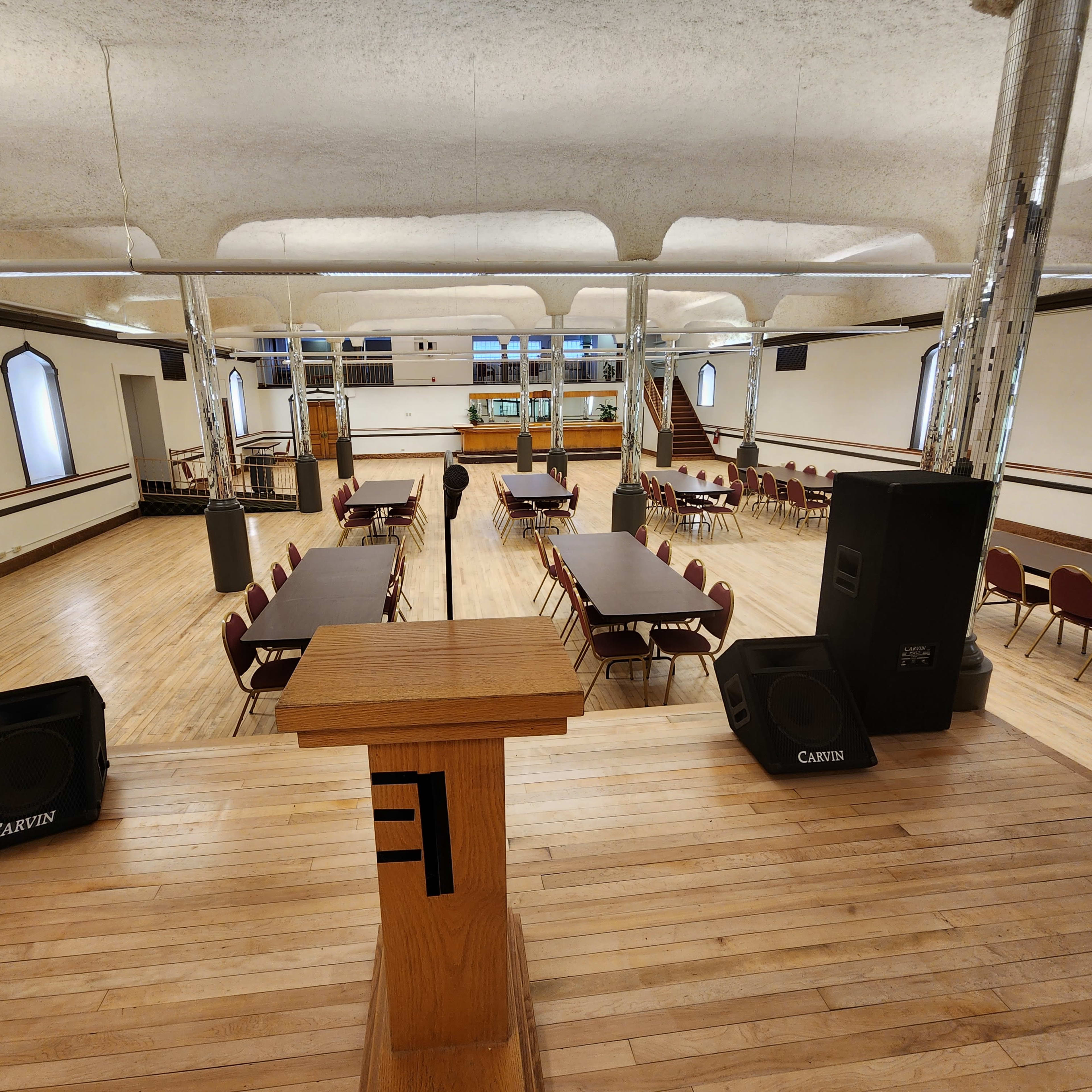 A spacious hall set up with several tables and chairs, featuring a podium at the front and sound equipment on either side.