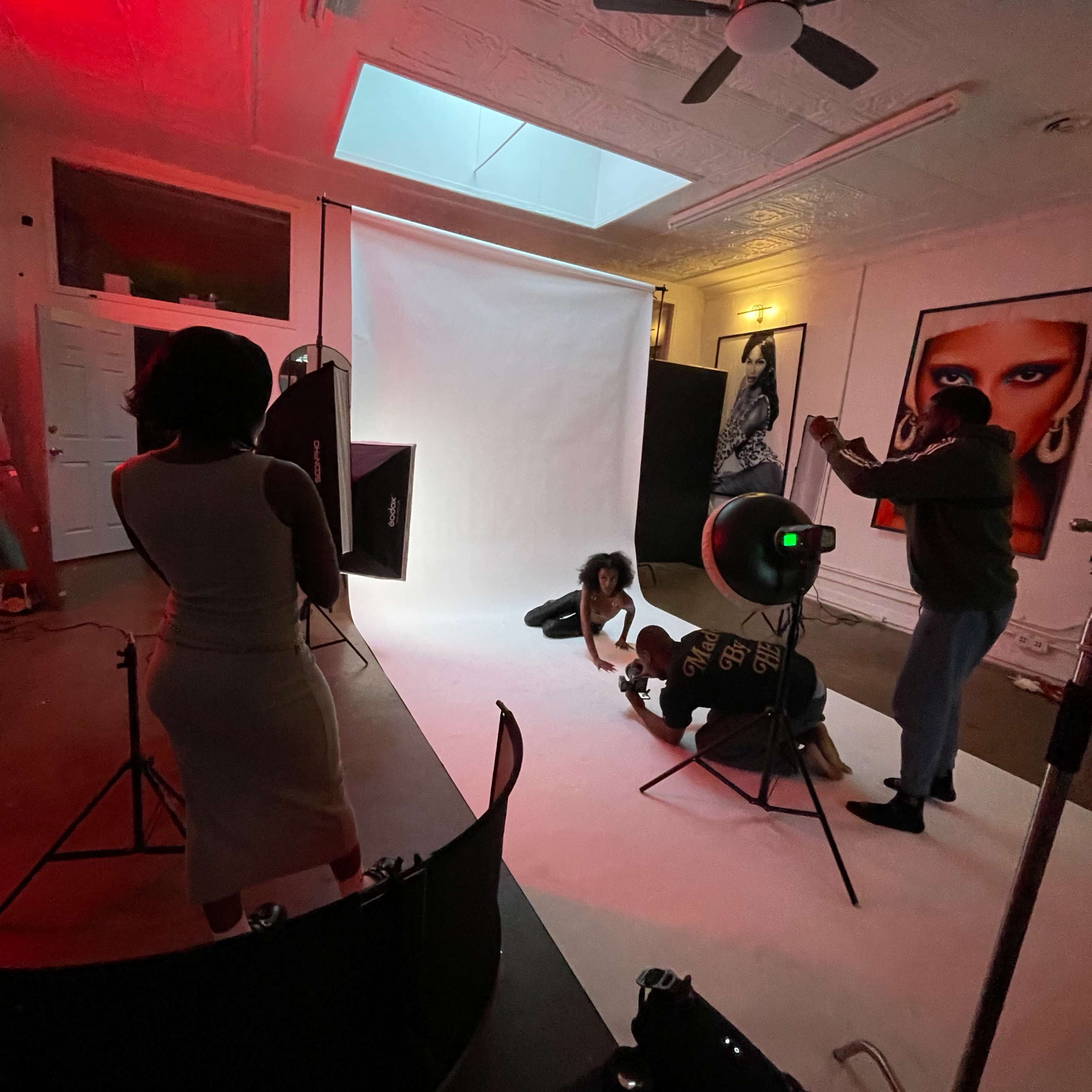 A photoshoot is taking place in a studio with a white backdrop and several lighting equipment setups, featuring a model on the floor and photographers adjusting their cameras.