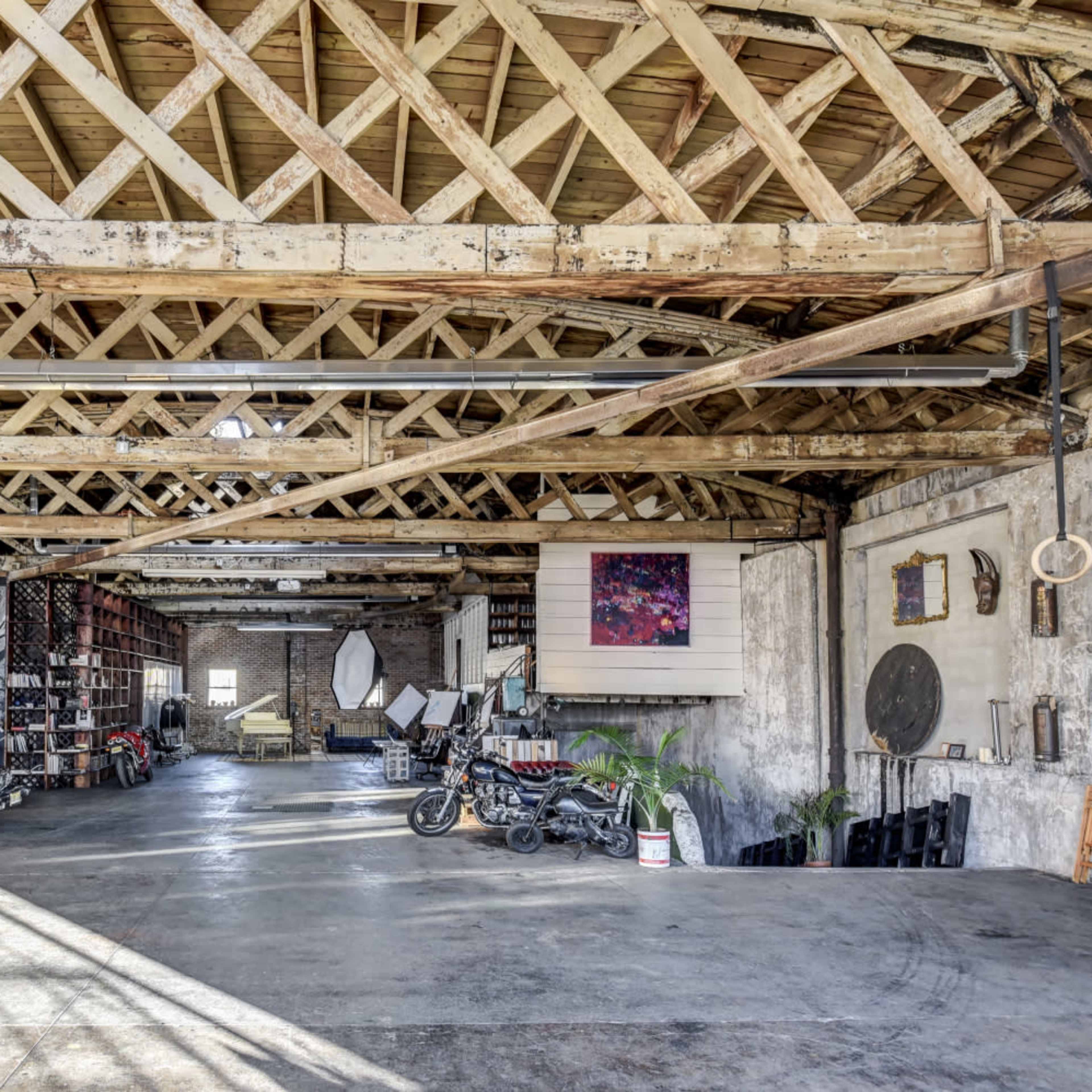 A spacious, industrial-style warehouse interior with wooden beams, various furniture items, a parked car, and artistic decorations on the walls.
