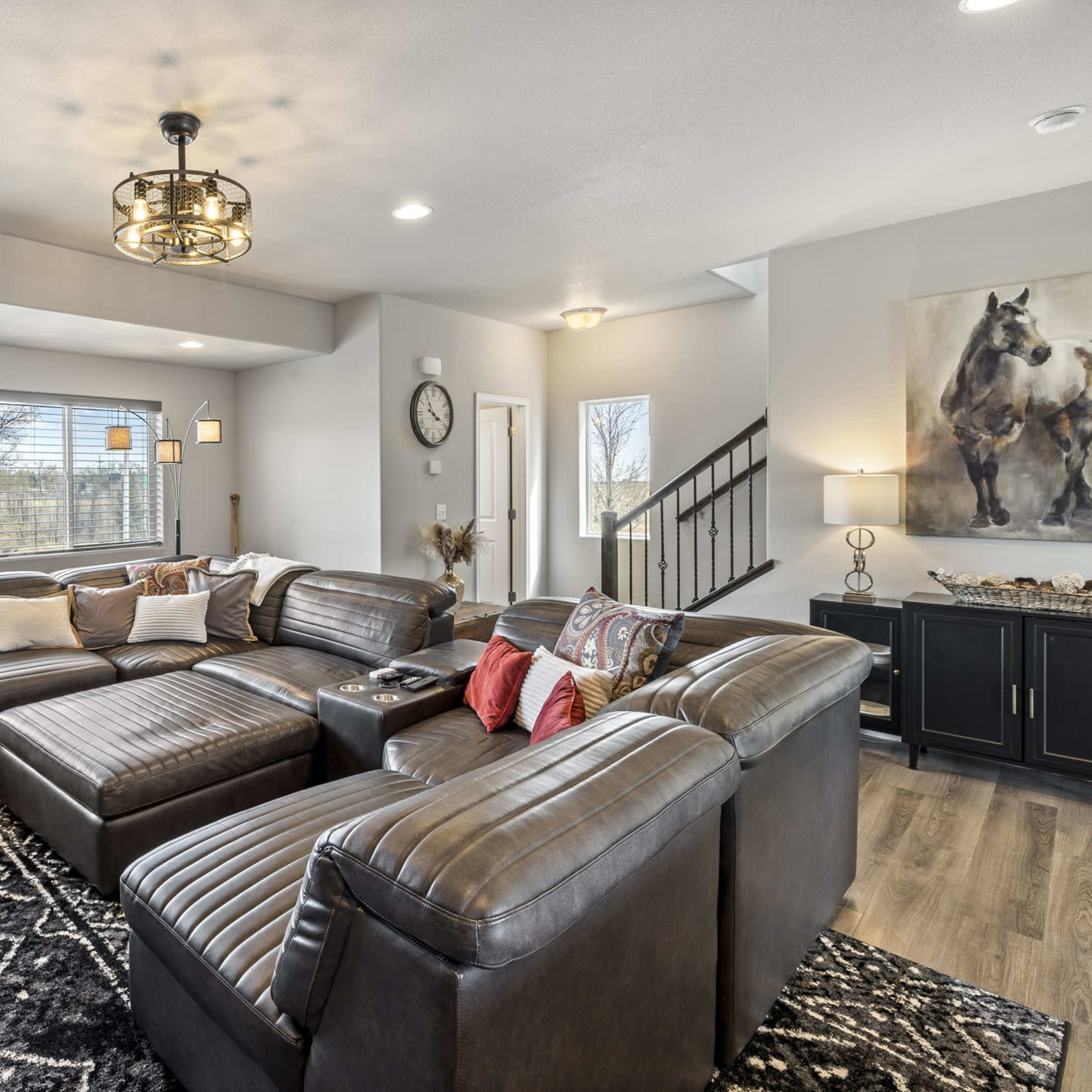 A spacious living room with a large, dark sectional sofa, a coffee table, and windows allowing natural light, featuring a modern chandelier and a painting of a horse on the wall.