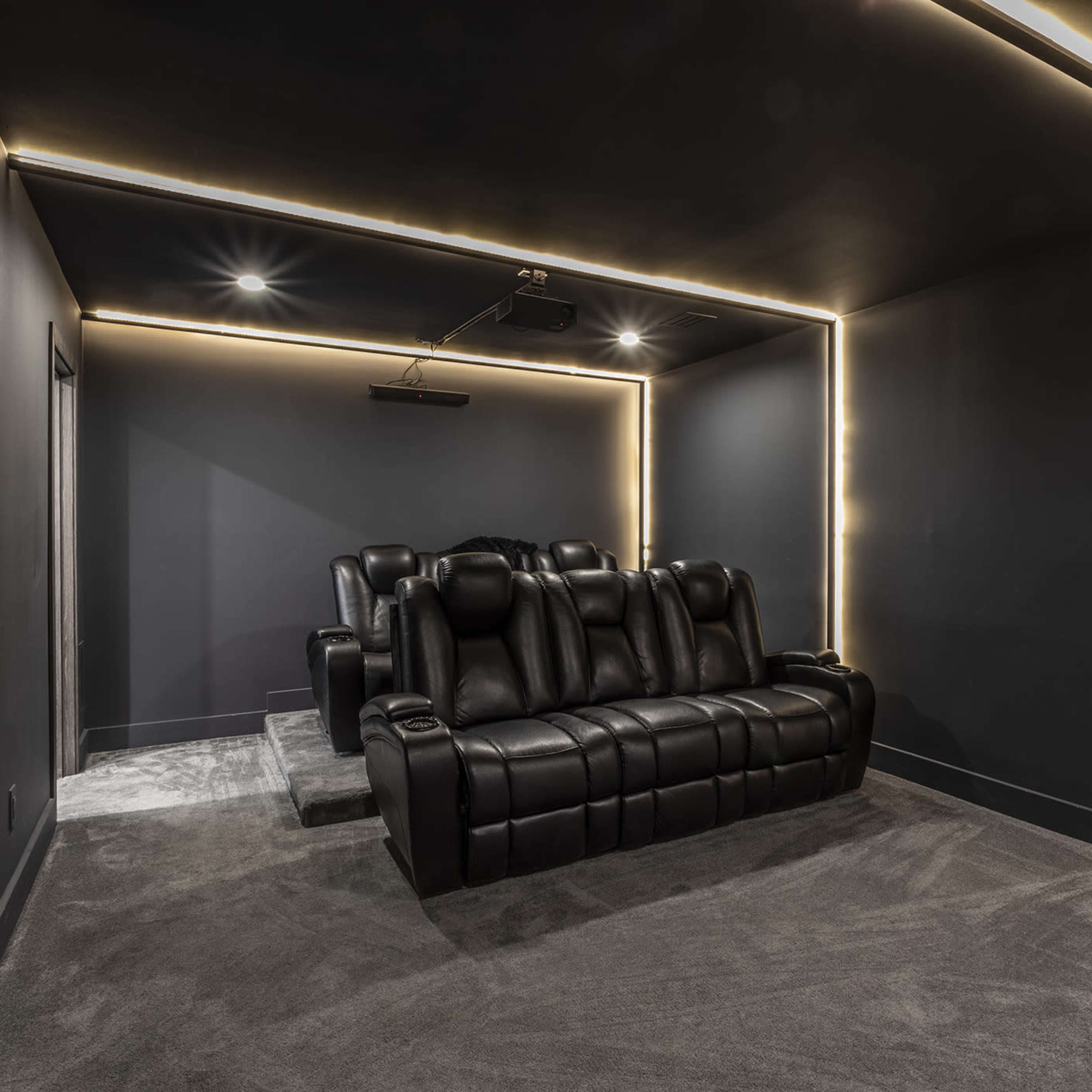 A dimly lit home theater room with black recliner seating and LED strip lighting along the walls.