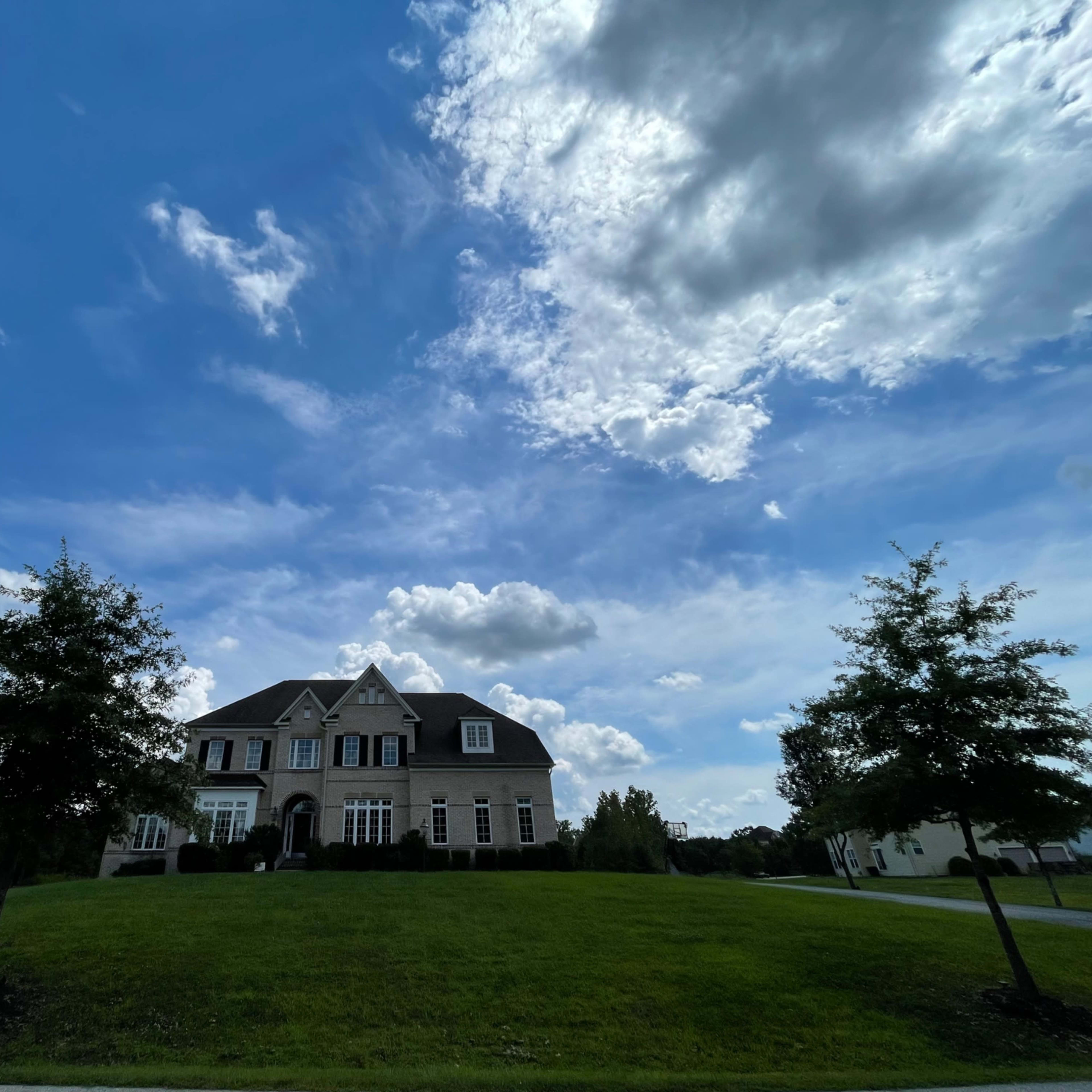 A large house with a manicured lawn is set against a backdrop of blue sky and scattered clouds.