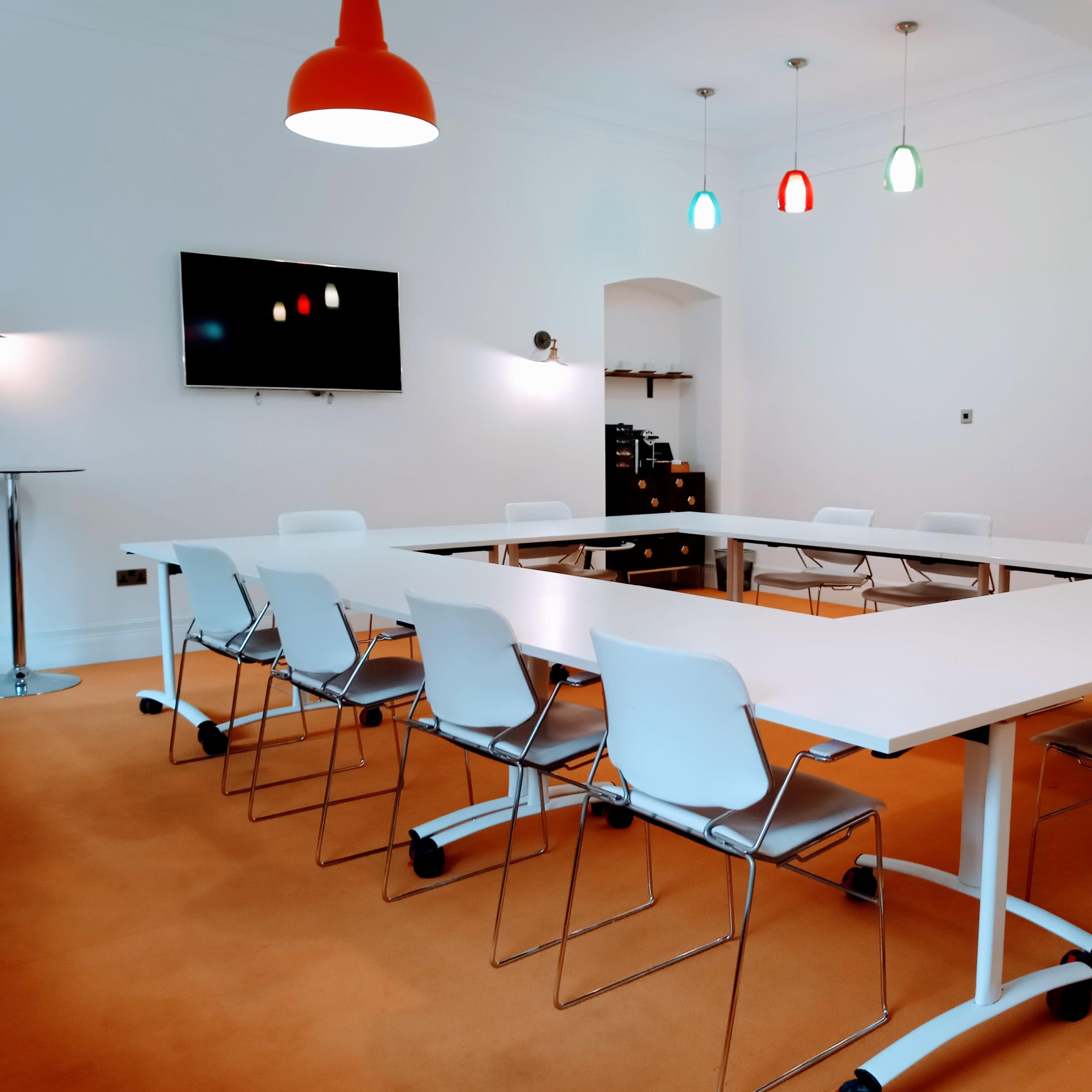 A modern conference room features a large white table surrounded by eight white chairs, with a television mounted on the wall and colorful pendant lights hanging from the ceiling.