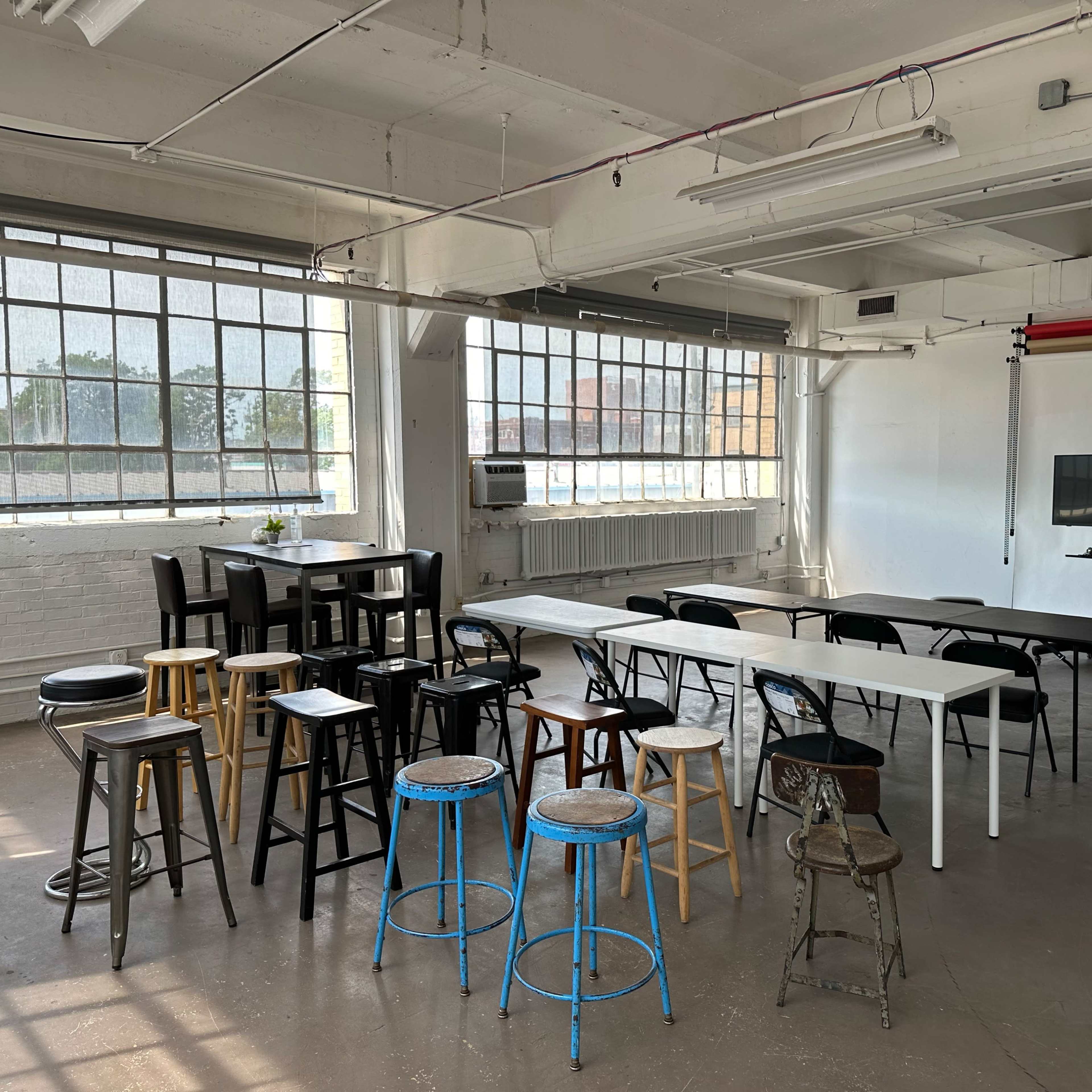 A spacious, bright room with large windows, furnished with various types of stools and tables arranged in different configurations.