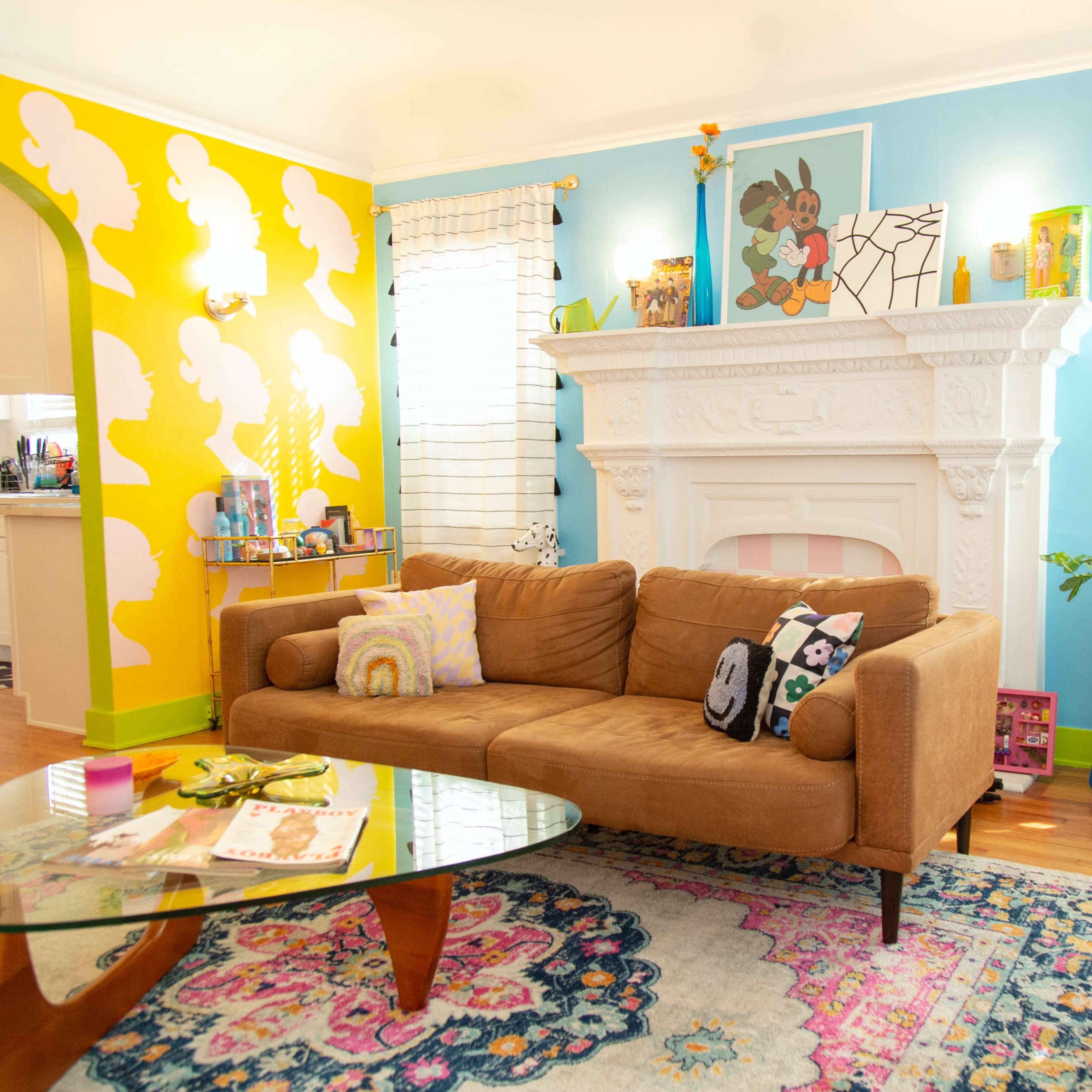 A brightly decorated living room with a brown sofa, a glass coffee table, colorful wall accents, and various decorative items arranged around a white fireplace.