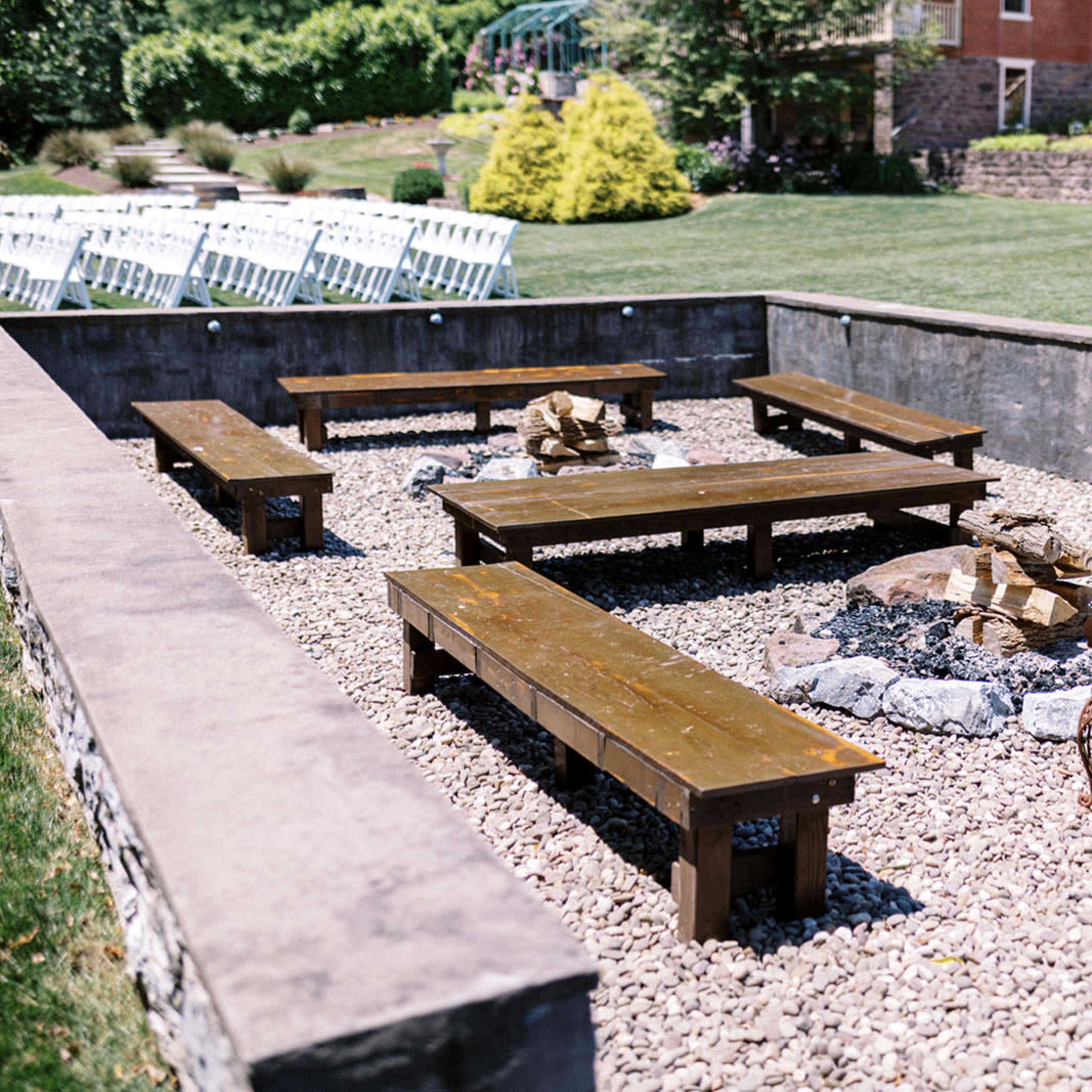 An outdoor fire pit area surrounded by wooden benches and wrought iron chairs, with a gravel surface and landscaping in the background.
