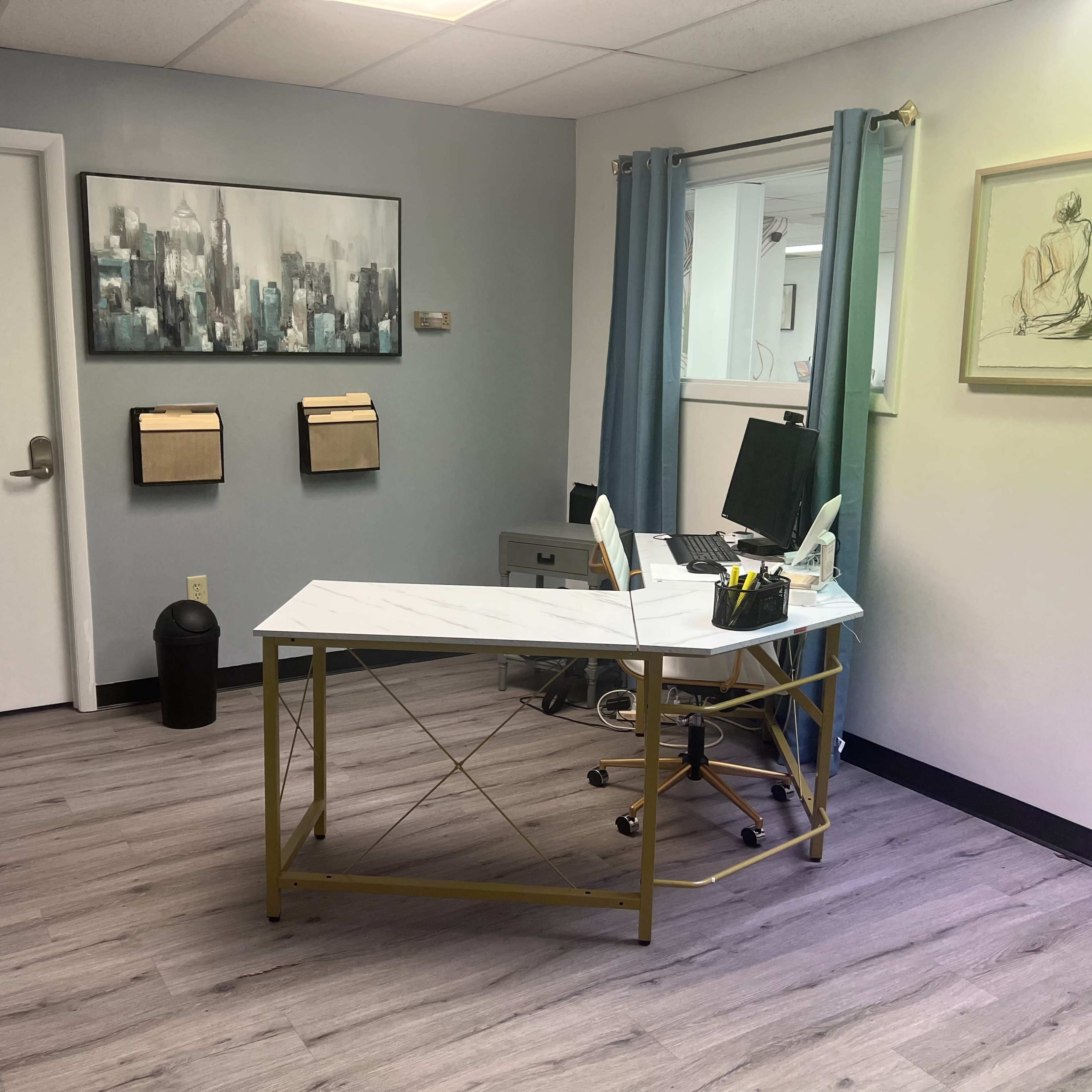 A modern office space featuring a white desk with gold legs, a computer chair, wall-mounted mail organizers, and artwork on the walls.