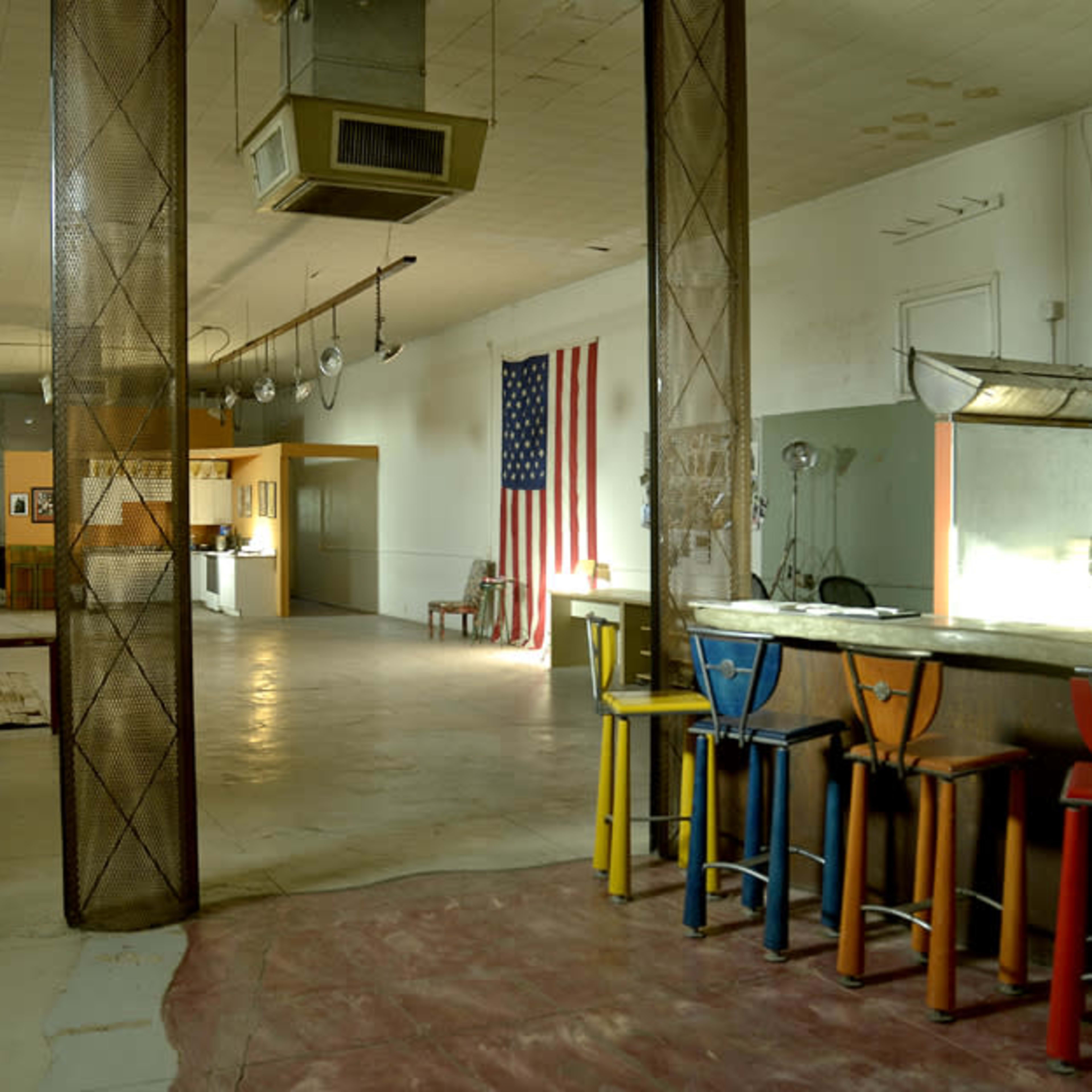 An interior space with a bar featuring colorful stools, an American flag on the wall, and various art pieces and furniture scattered throughout the room.