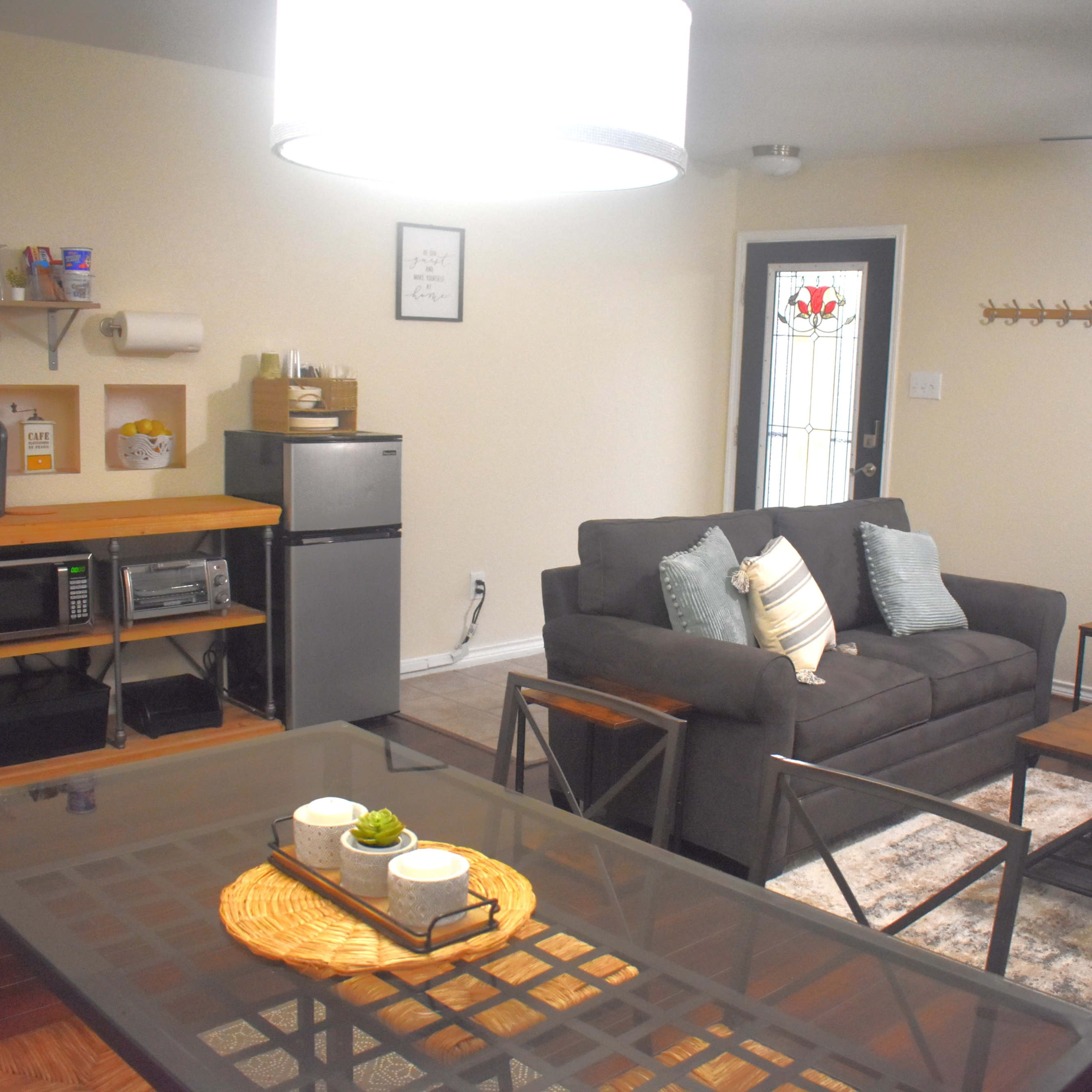 A cozy living space with a dining area, a gray sofa, a blue armchair, a small kitchen setup with appliances, and a door leading outside.