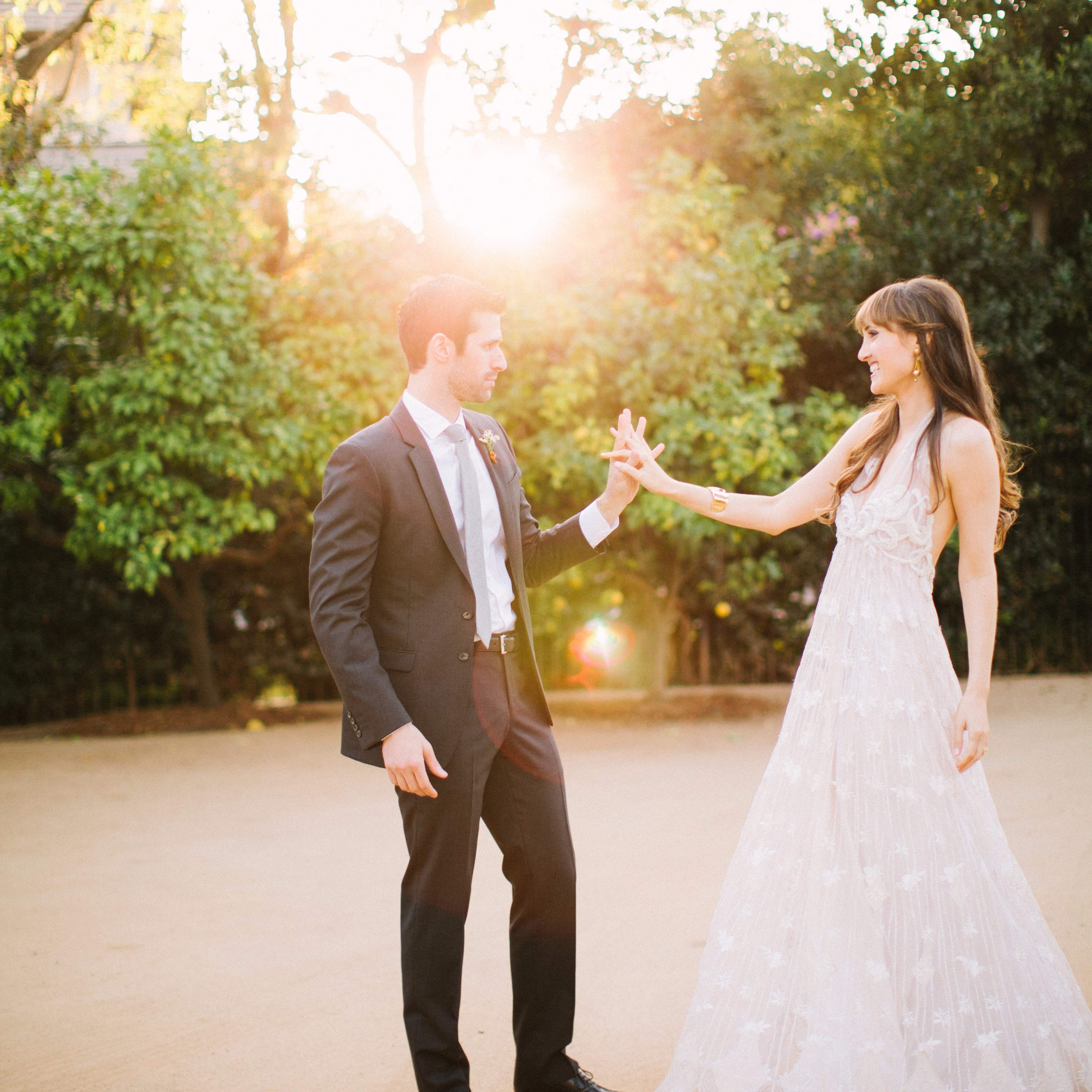 A couple stands on a dirt path in a garden, holding hands as the sun sets behind them.