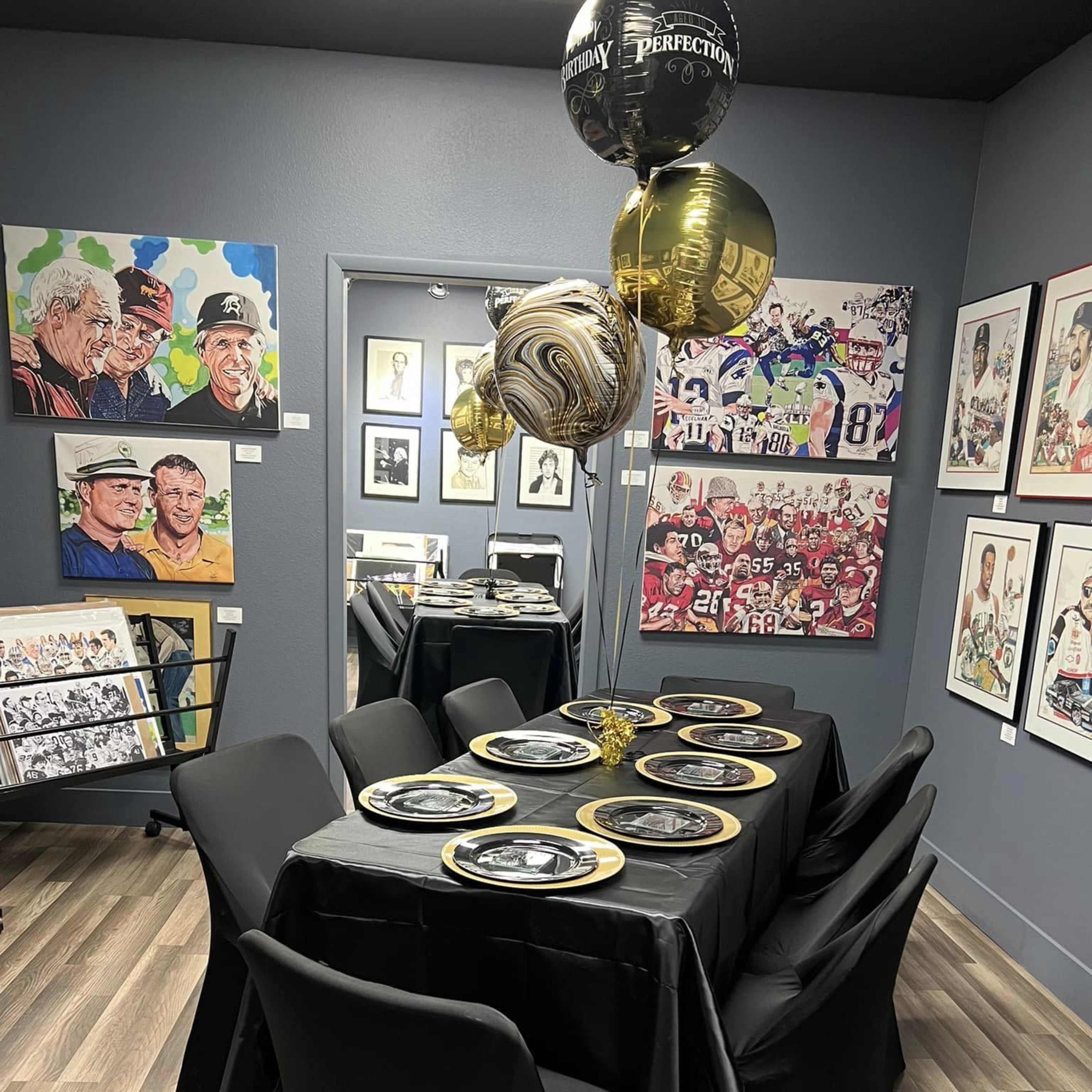 A decorated dining area with a black tablecloth-covered table set for a meal, featuring gold and black balloons and framed artwork on the walls.