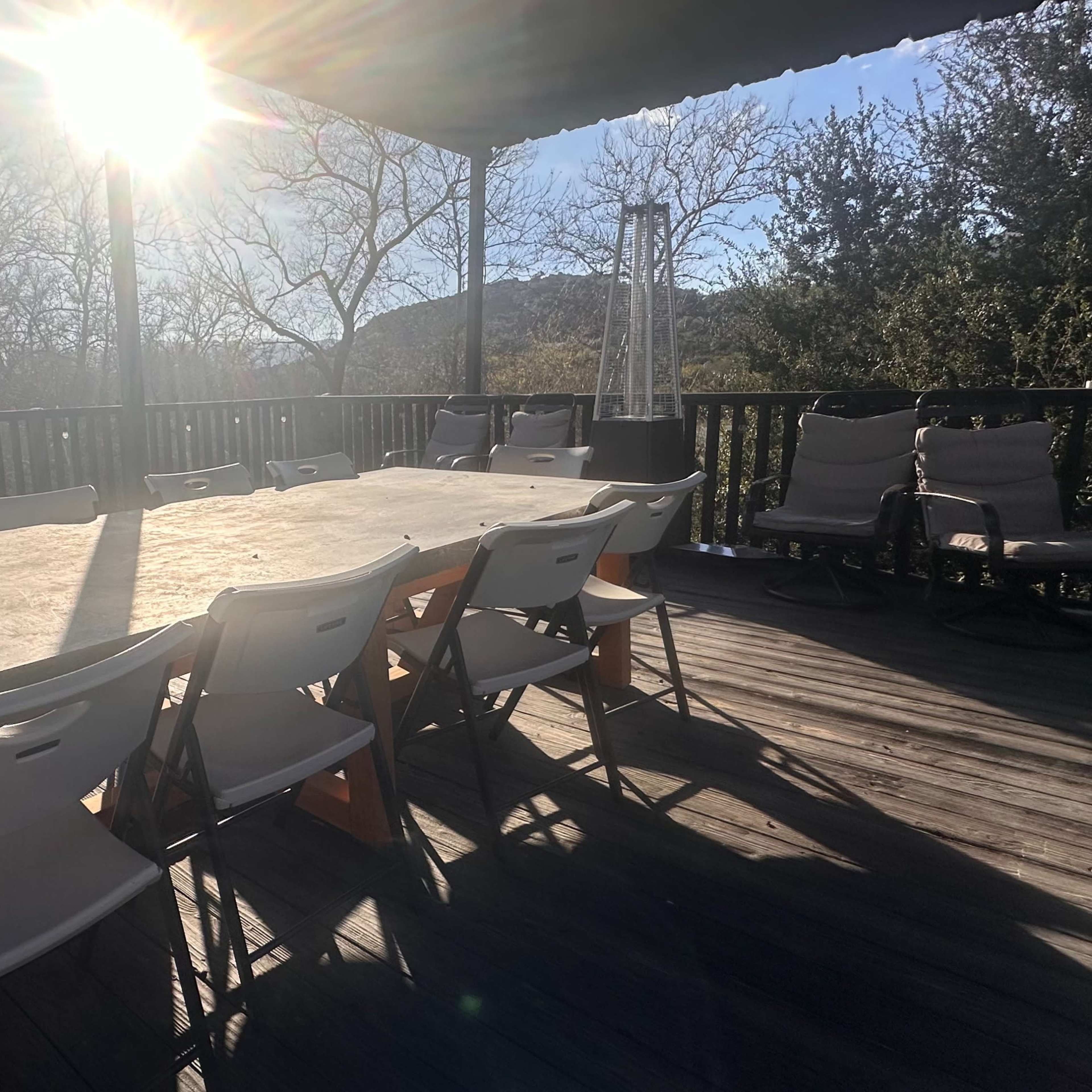 A long wooden table with white chairs is set on a deck under bright sunlight, surrounded by trees and hills in the background.