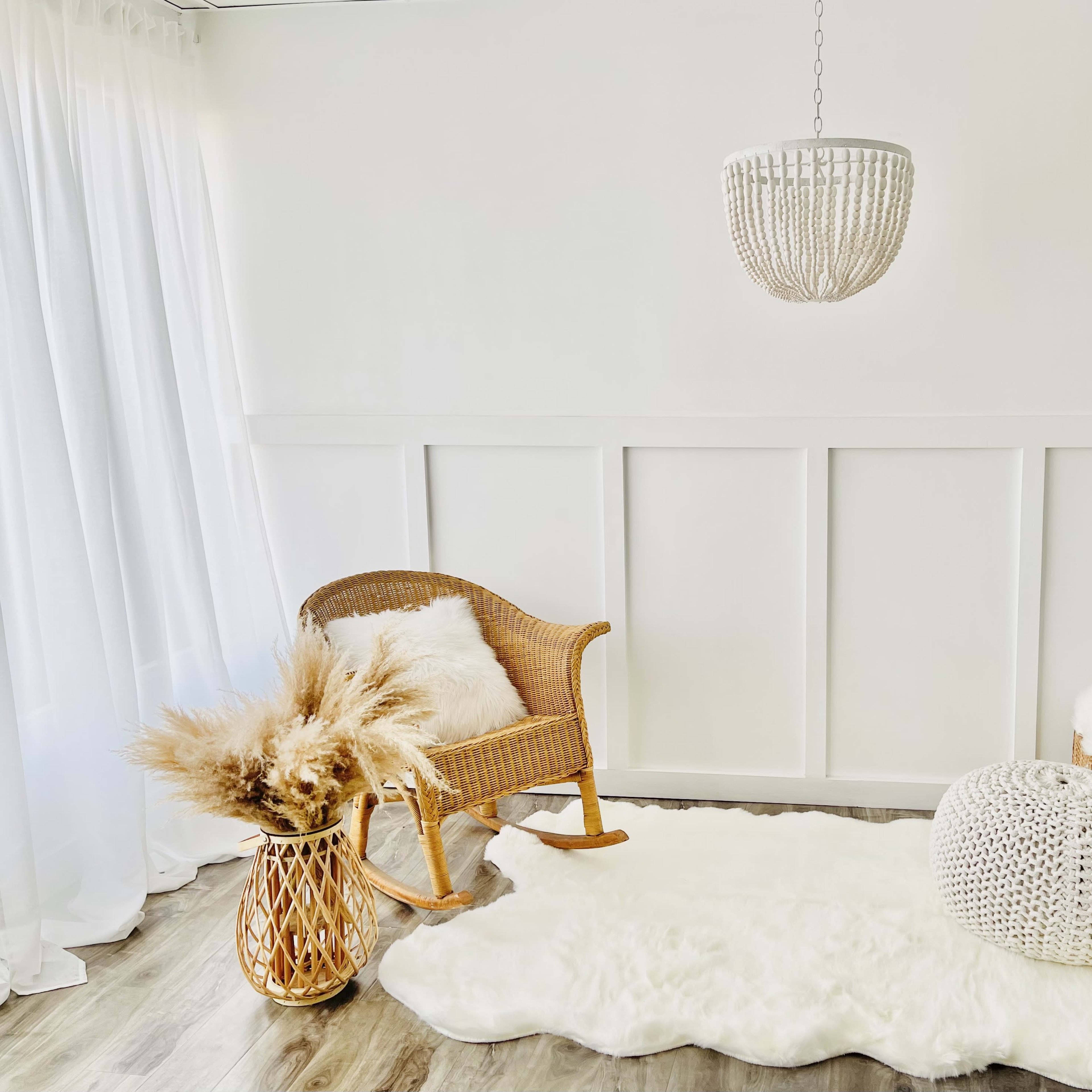 A cozy interior corner with a wicker rocking chair, a hanging light fixture, a plush white rug, a decorative pouf, and a basket holding pampas grass.