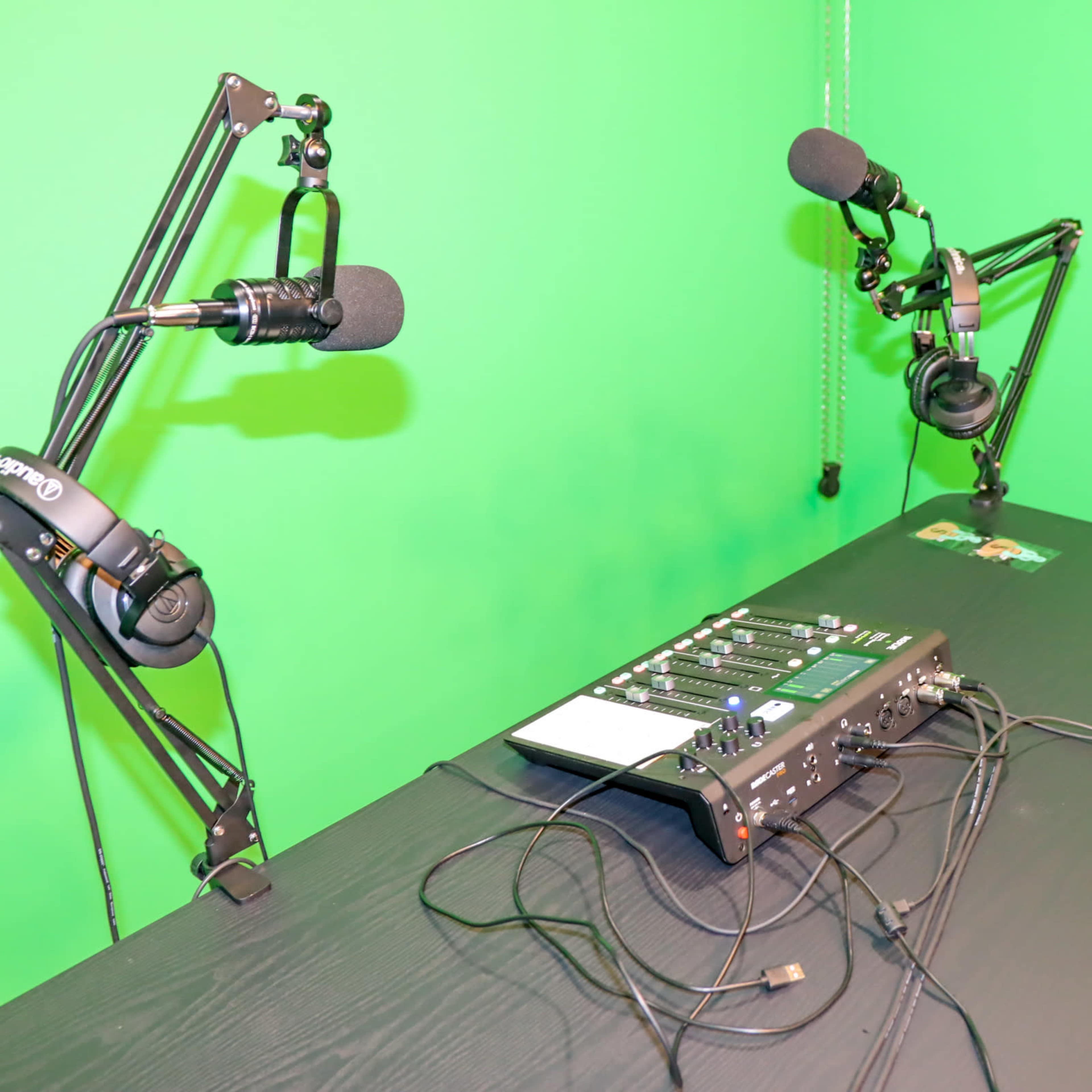 A black table with microphones mounted on adjustable arms and an audio mixer is placed in front of a green screen.
