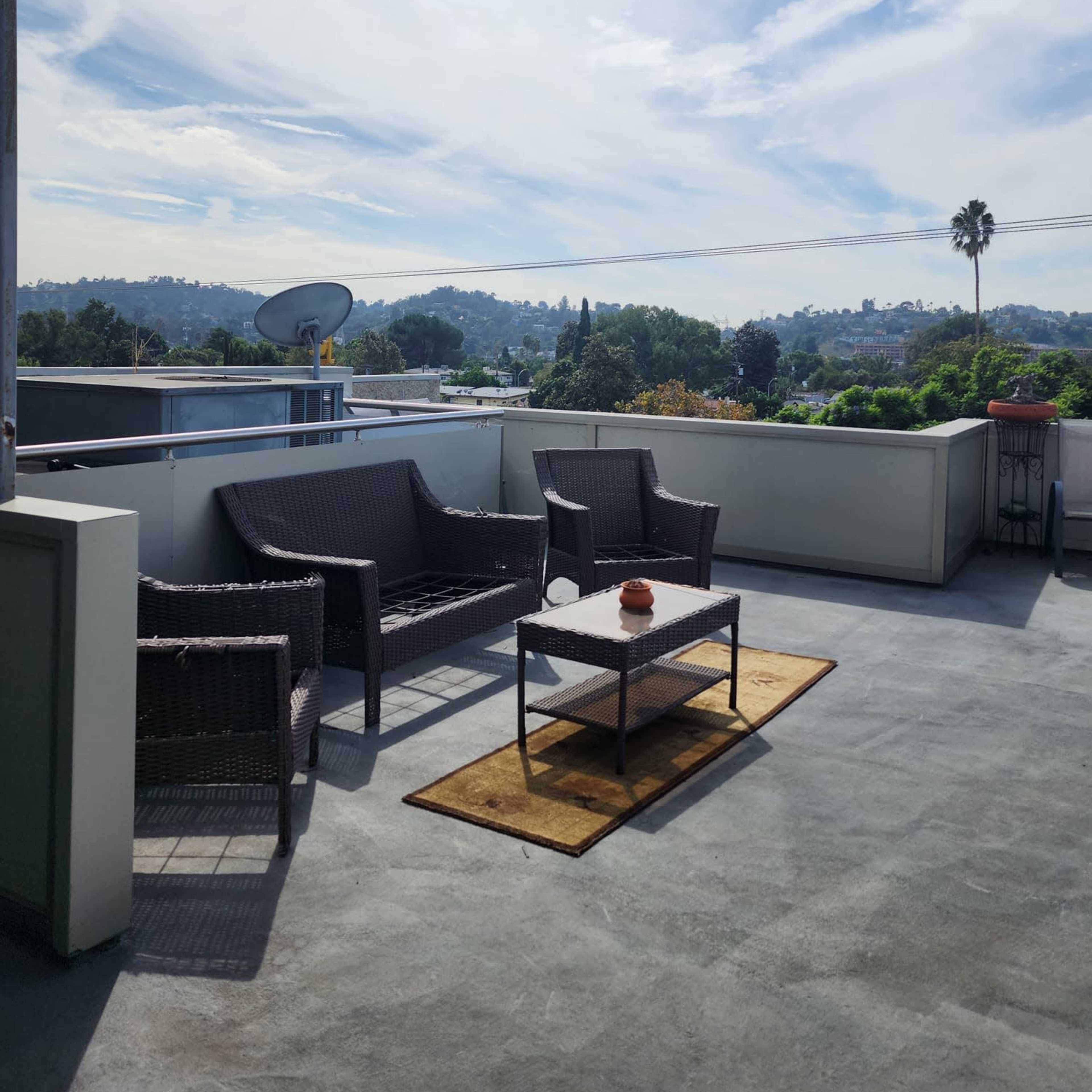 A modern outdoor terrace features wicker furniture, a table with a small centerpiece, and views of hills and trees under a partly cloudy sky.