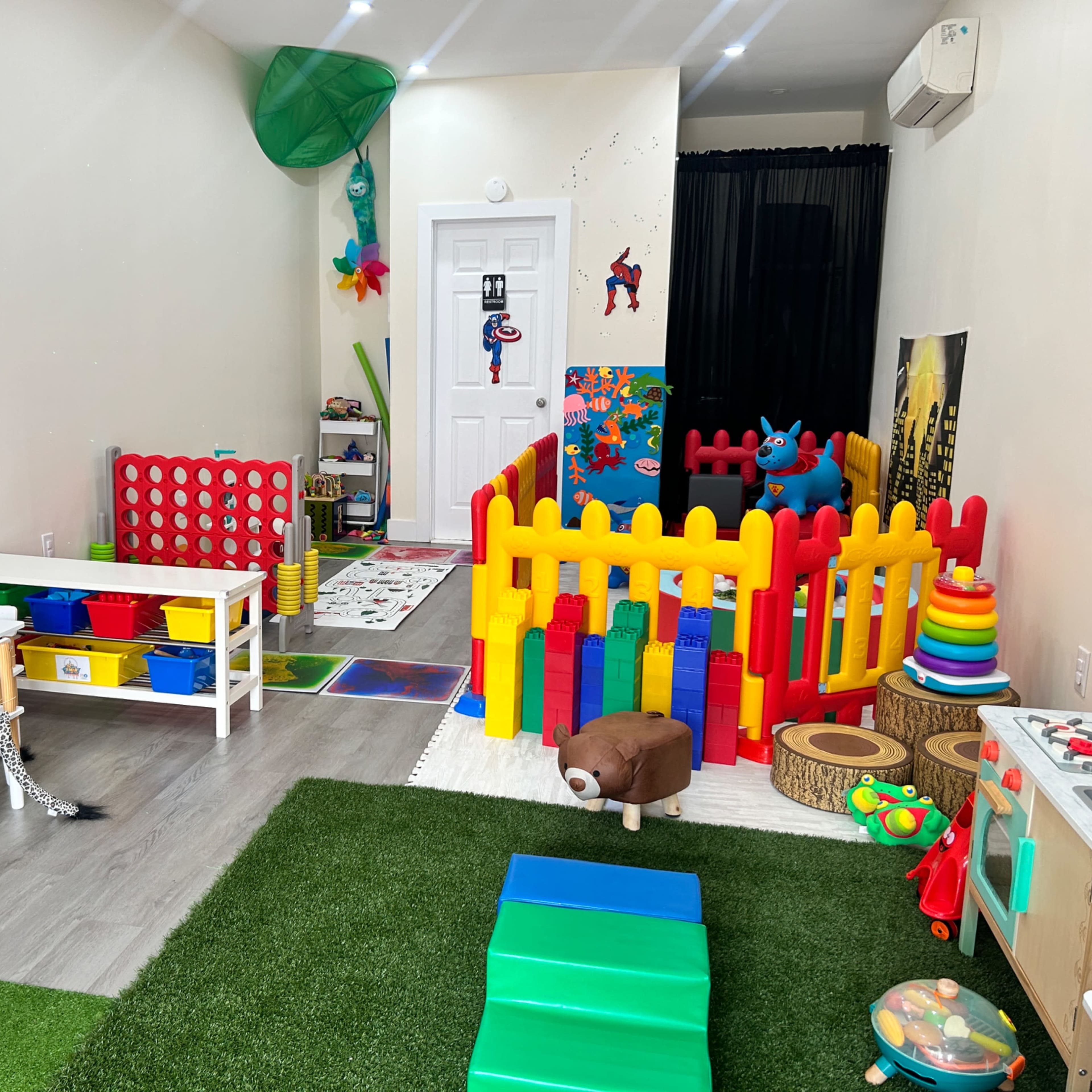 A colorful playroom filled with various children's toys, including a fenced play area, tables, and a green carpet.
