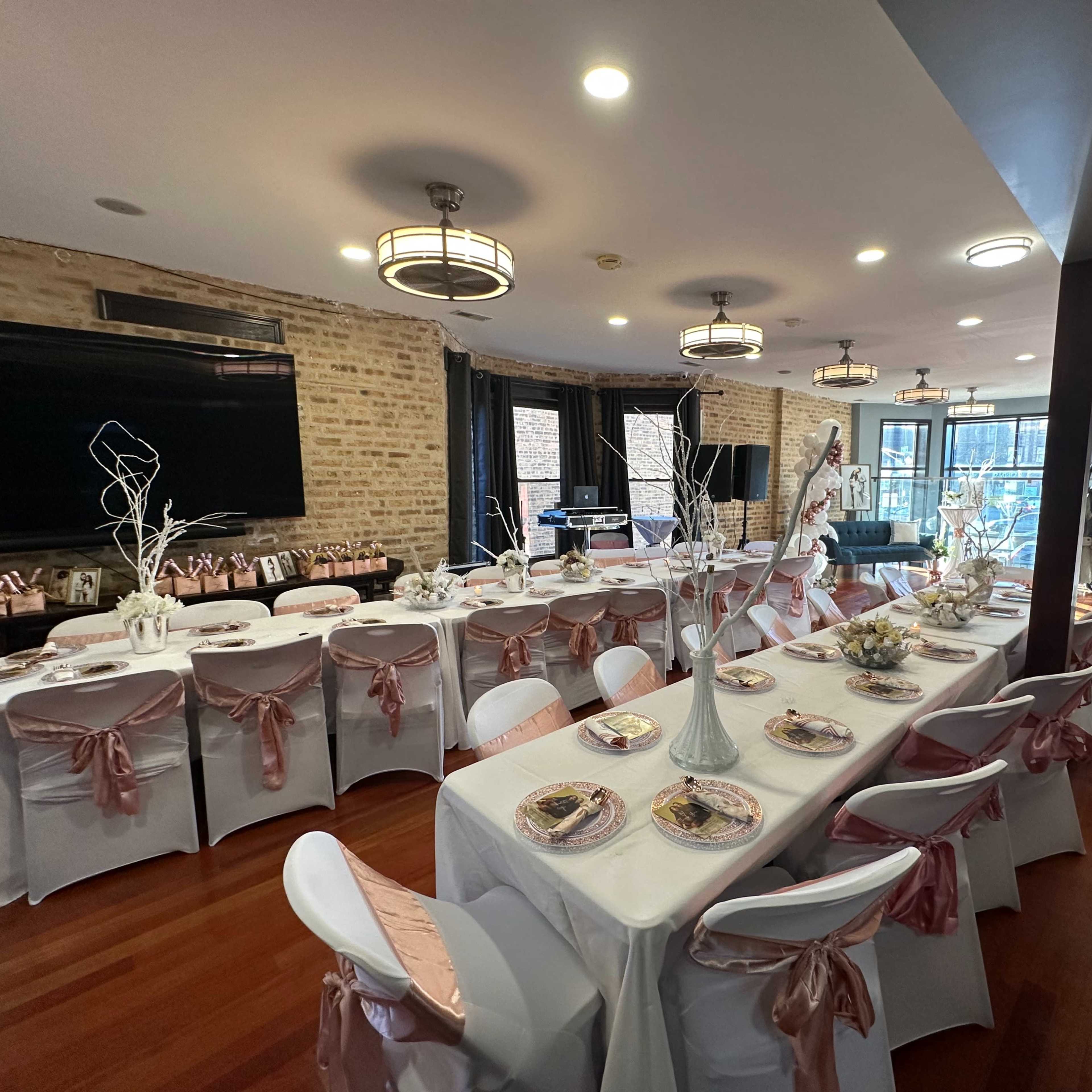 A dining area set for an event, featuring long tables adorned with white tablecloths, pink satin chair sashes, and decorative centerpieces.
