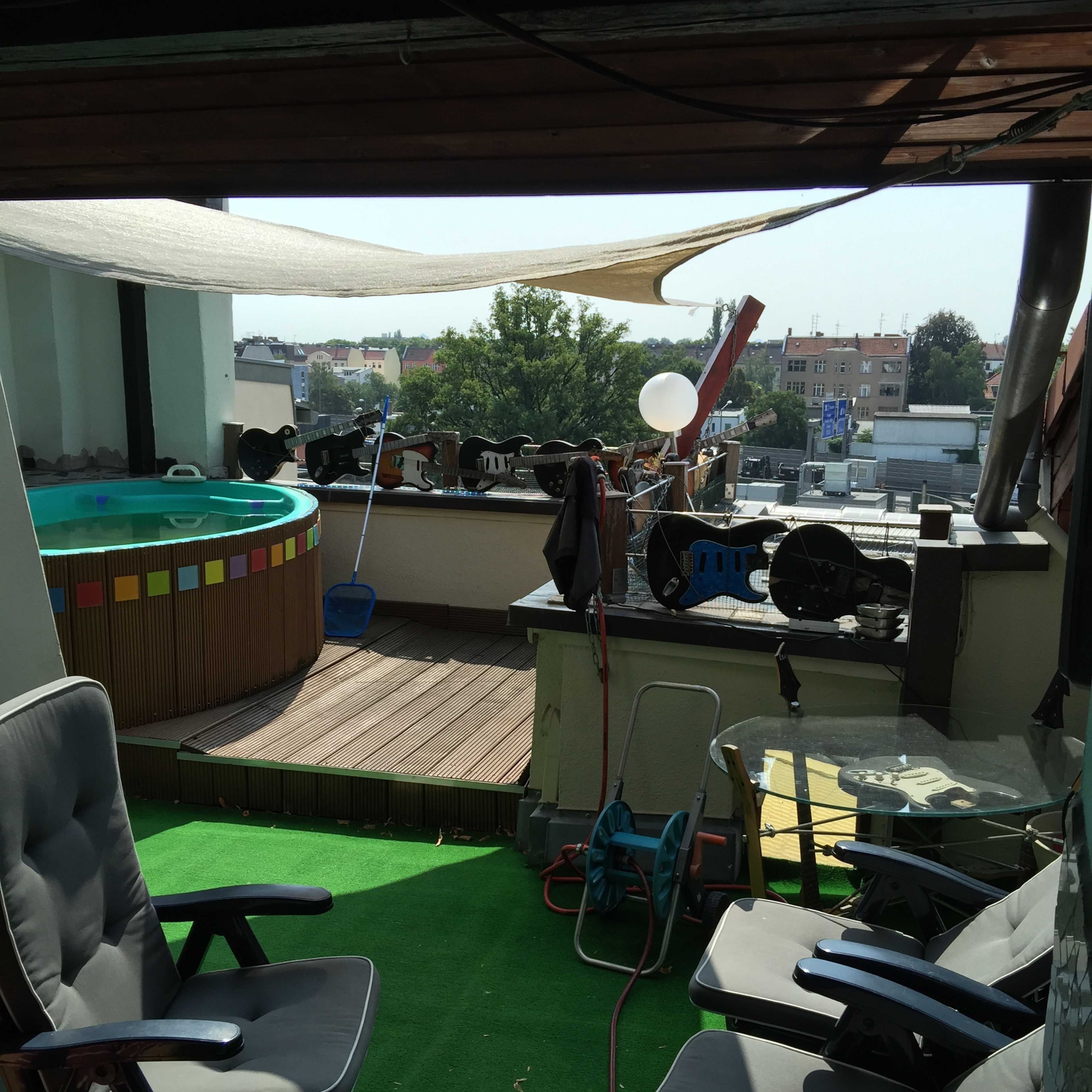 A rooftop terrace featuring a circular pool, lounge chairs, and a green carpeted floor, with a city view in the background.