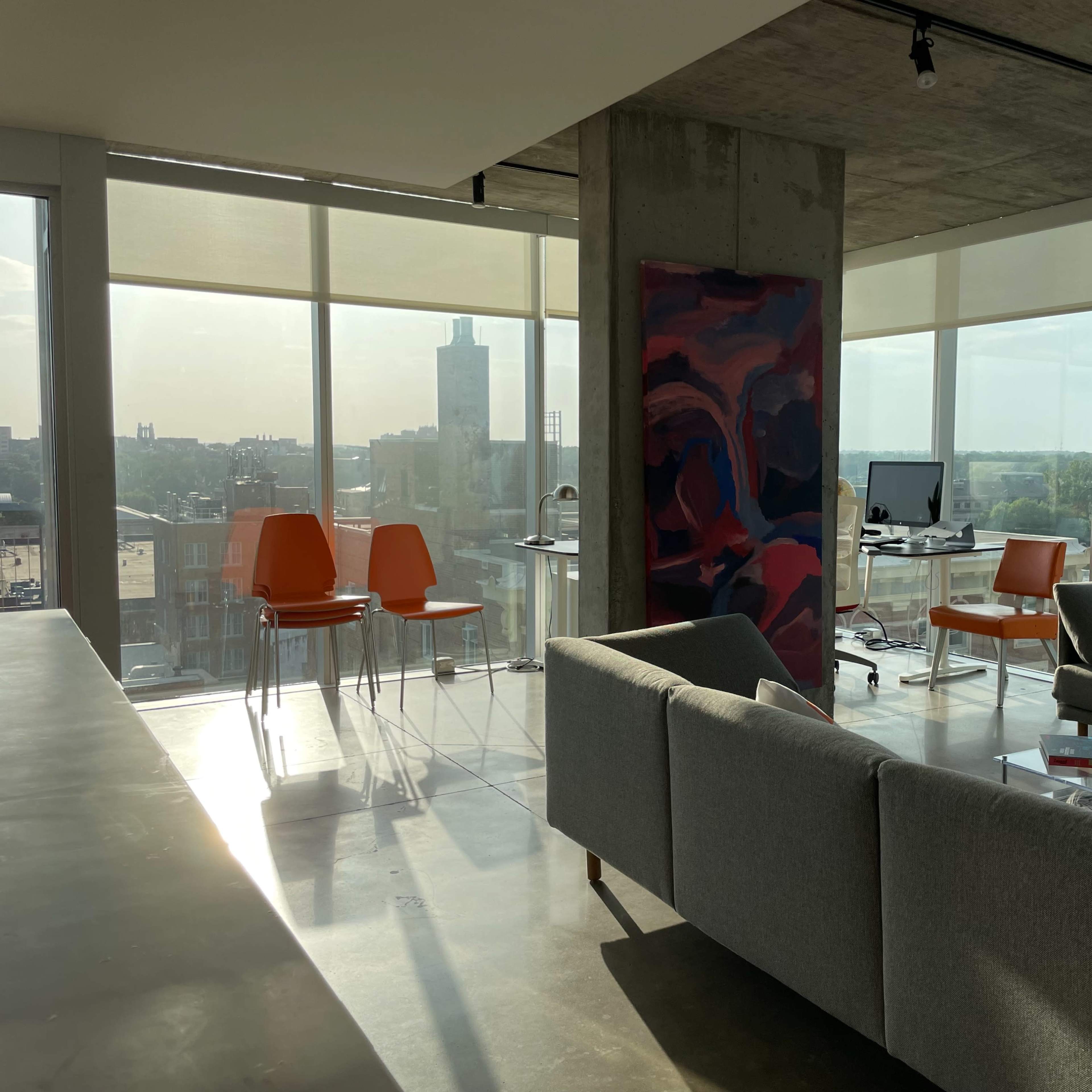 An open office space features a gray couch, orange chairs, a desk with a computer, and large windows that offer a view of the city.
