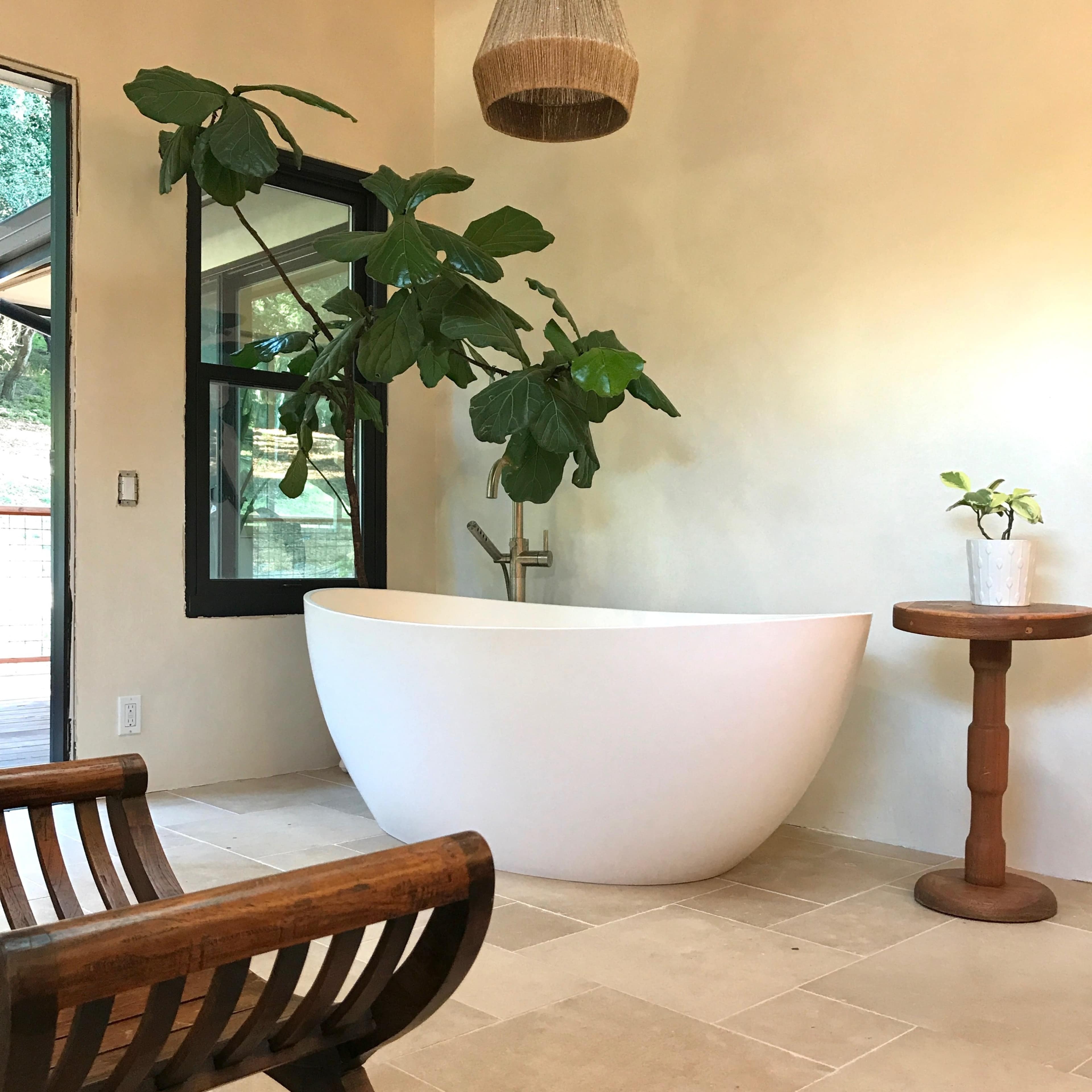 A modern bathroom featuring a freestanding white bathtub, a wooden chair, a small side table, and large indoor plants.
