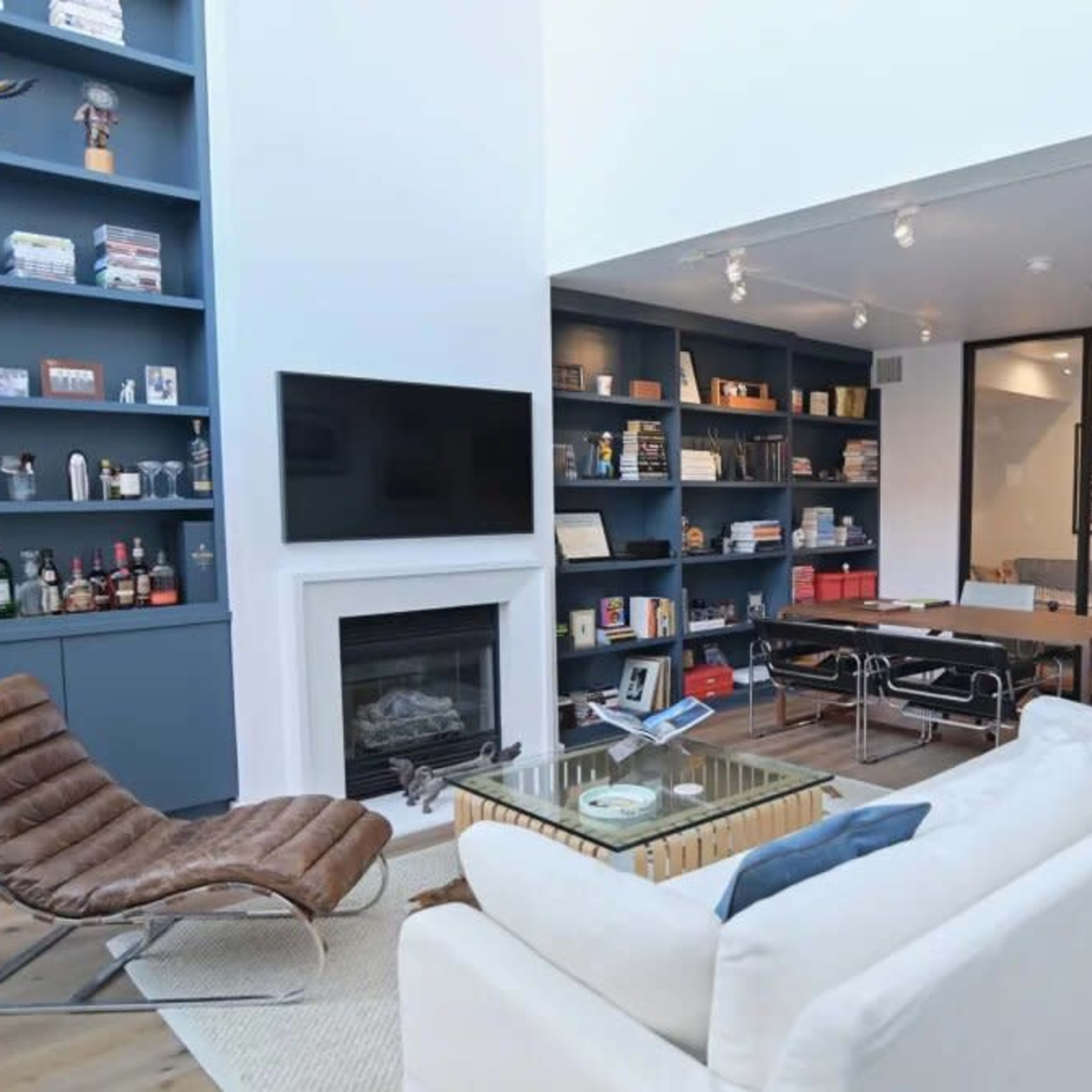 A modern living room with a white sofa, a glass coffee table, a fireplace, a wall-mounted television, and a bookshelf filled with books and decor items, leading into a dining area with a large wooden table.