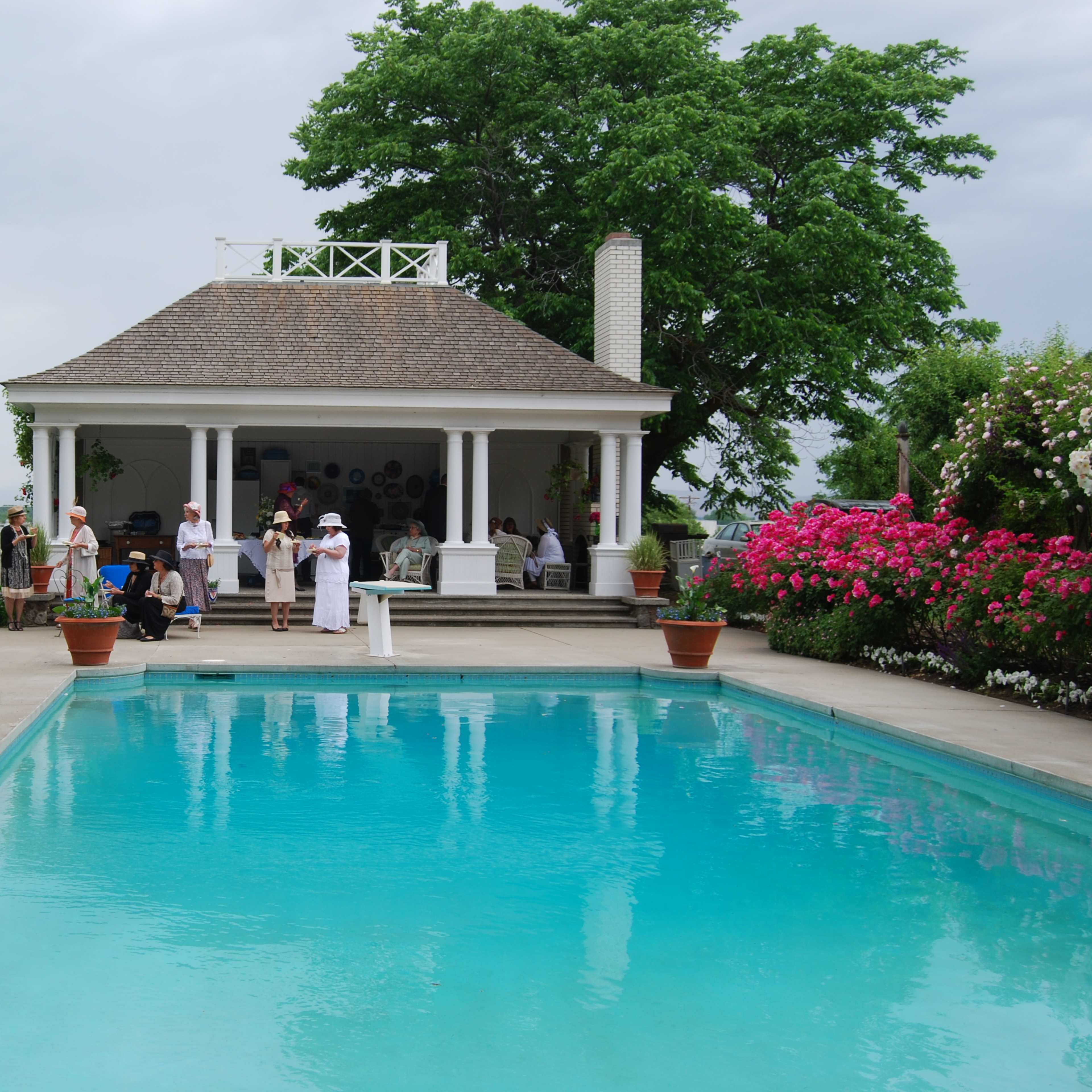 A gathering of people in formal attire attends an outdoor event by a clear swimming pool, surrounded by well-maintained gardens and a gazebo.