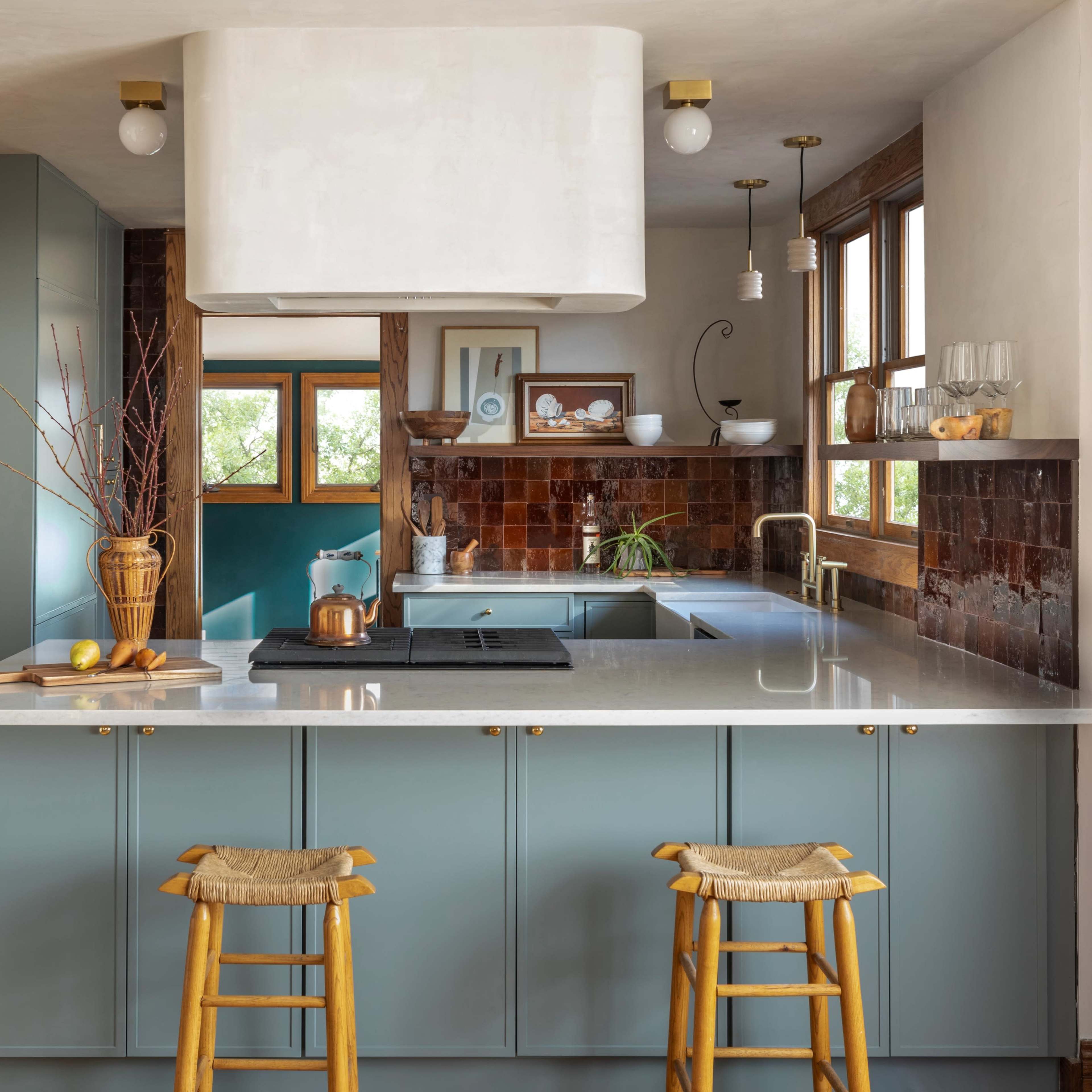 A modern kitchen features a central island with bar stools, green cabinetry, a large range hood, and warm-toned tile accents.
