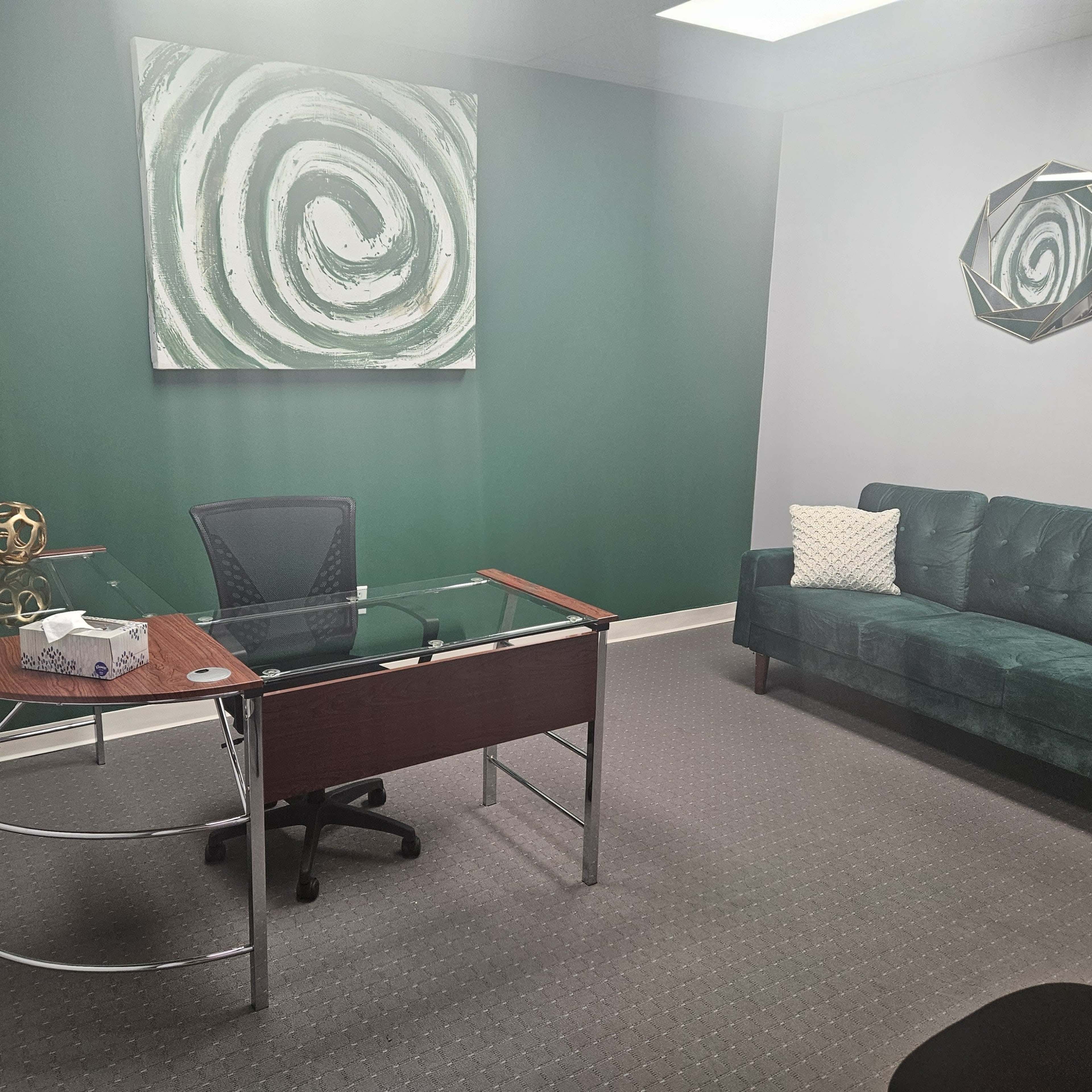 An office with a glass-top desk, a green couch, and a green spiral painting on the wall.