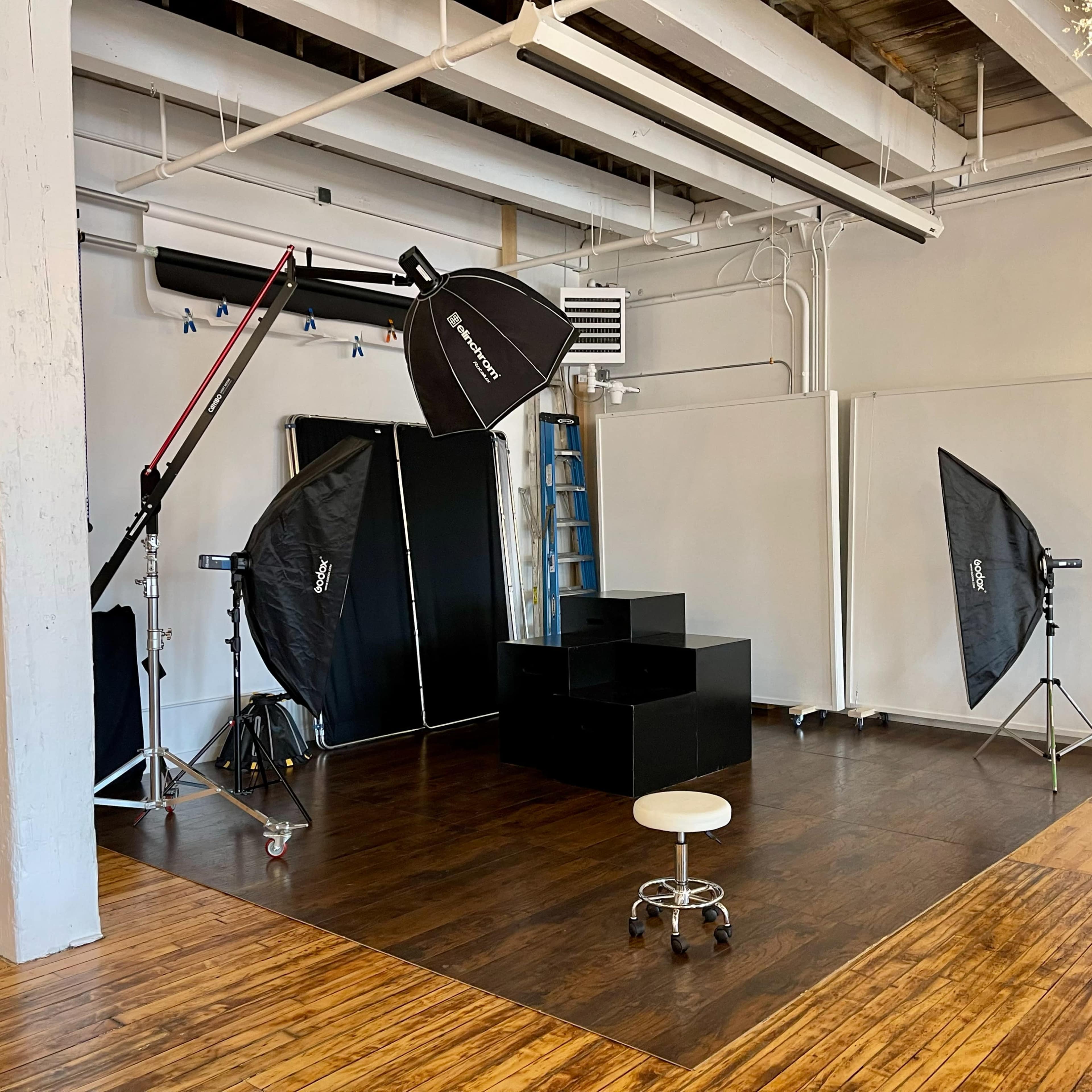 A photography studio setup with two softbox lights, a black backdrop, a stool, and a wooden floor.