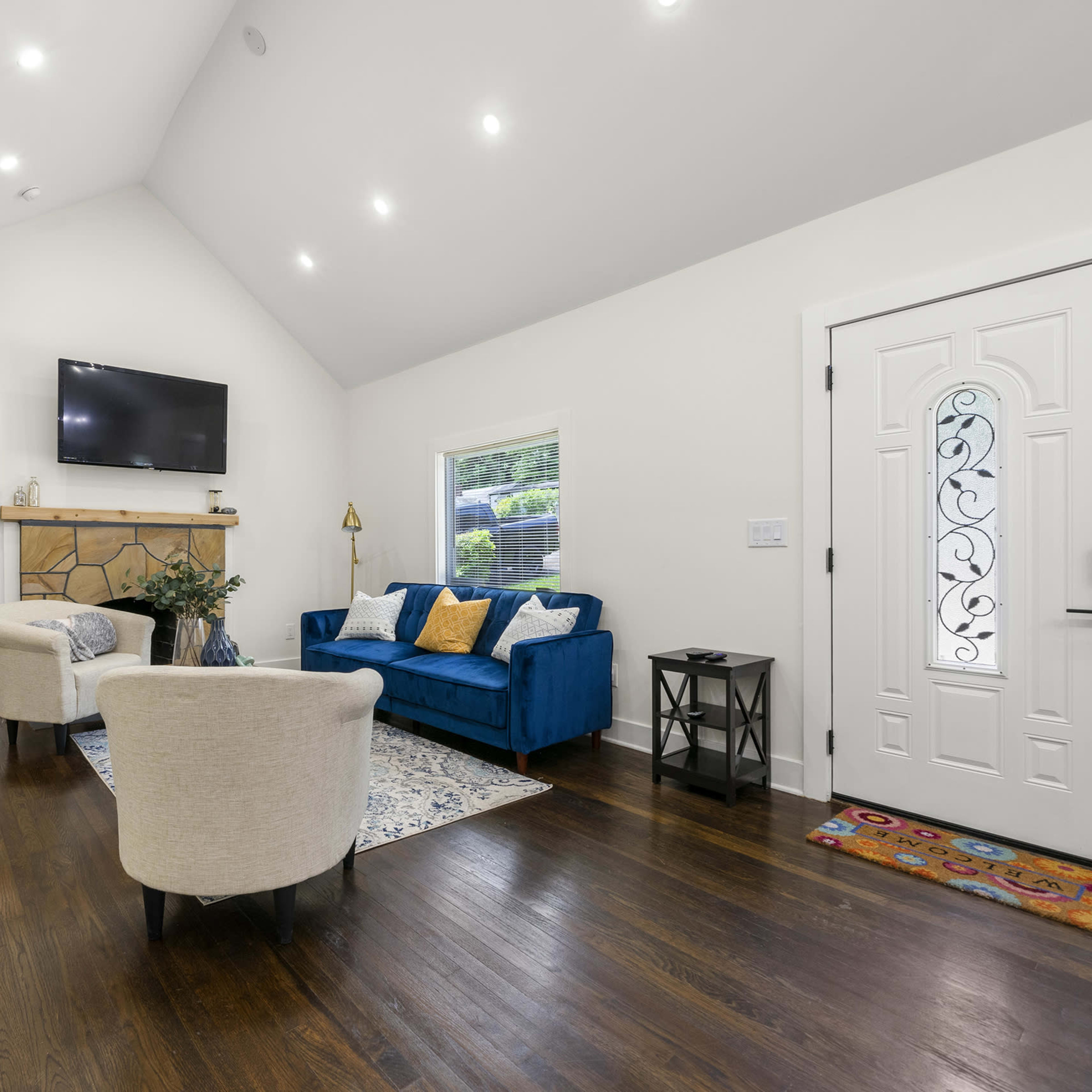 A living room featuring a blue sofa, two armchairs, a coffee table, a decorative rug, and a television mounted above a stone fireplace with a front door visible.