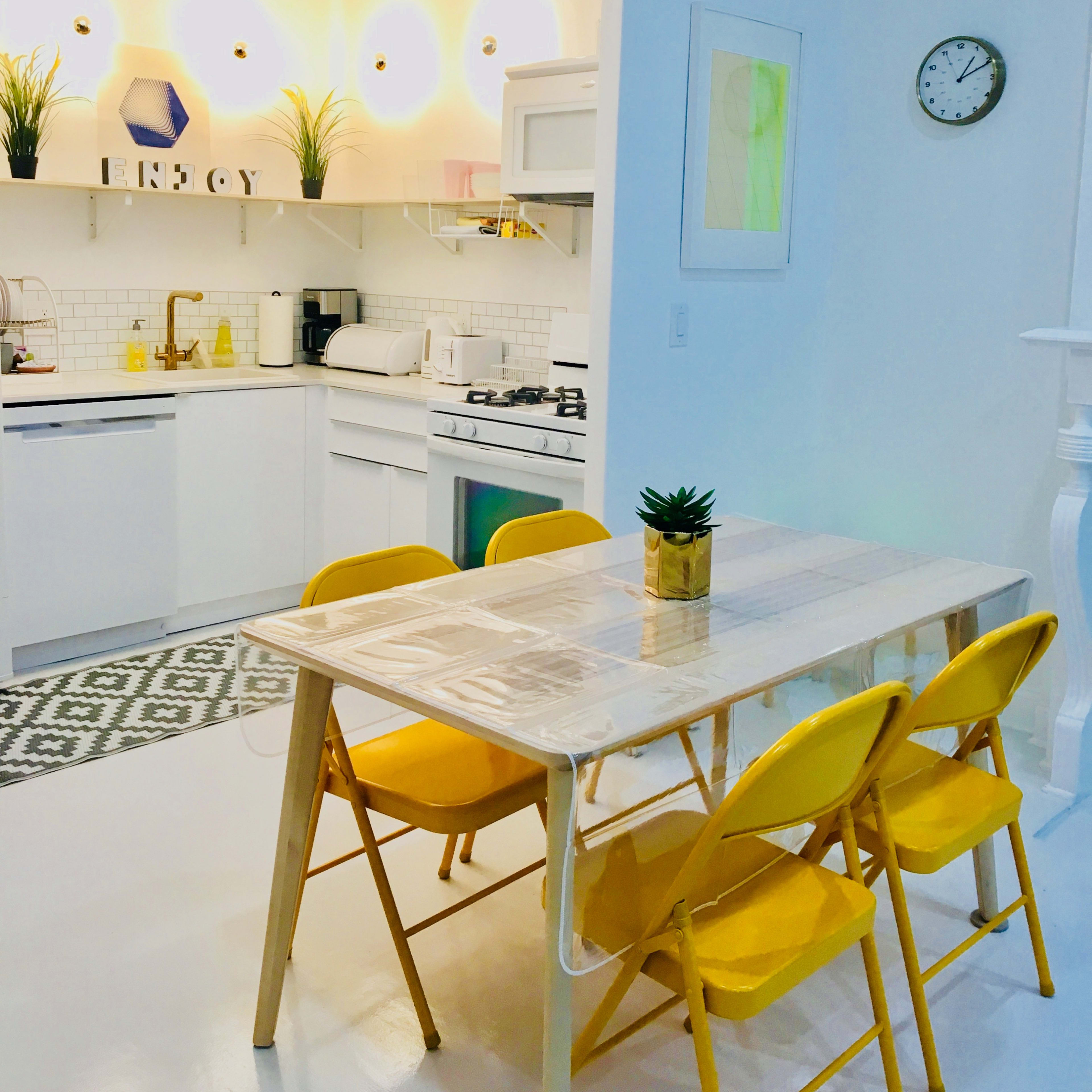 A bright, modern kitchen features a dining table with yellow chairs and a view of white cabinetry, plants, and a wall clock.