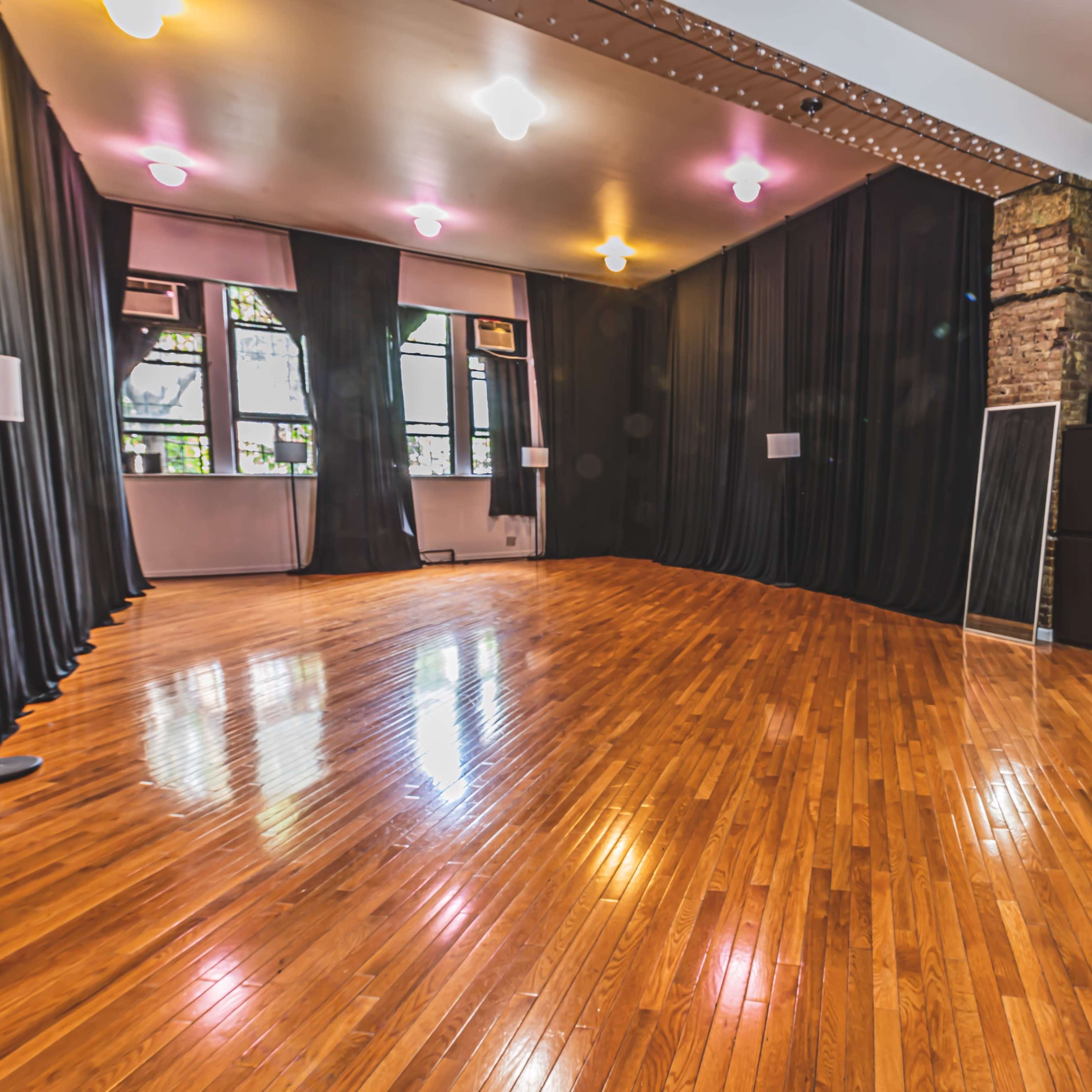 A spacious wooden-floored room with brick walls, large windows, and black curtains on two sides.