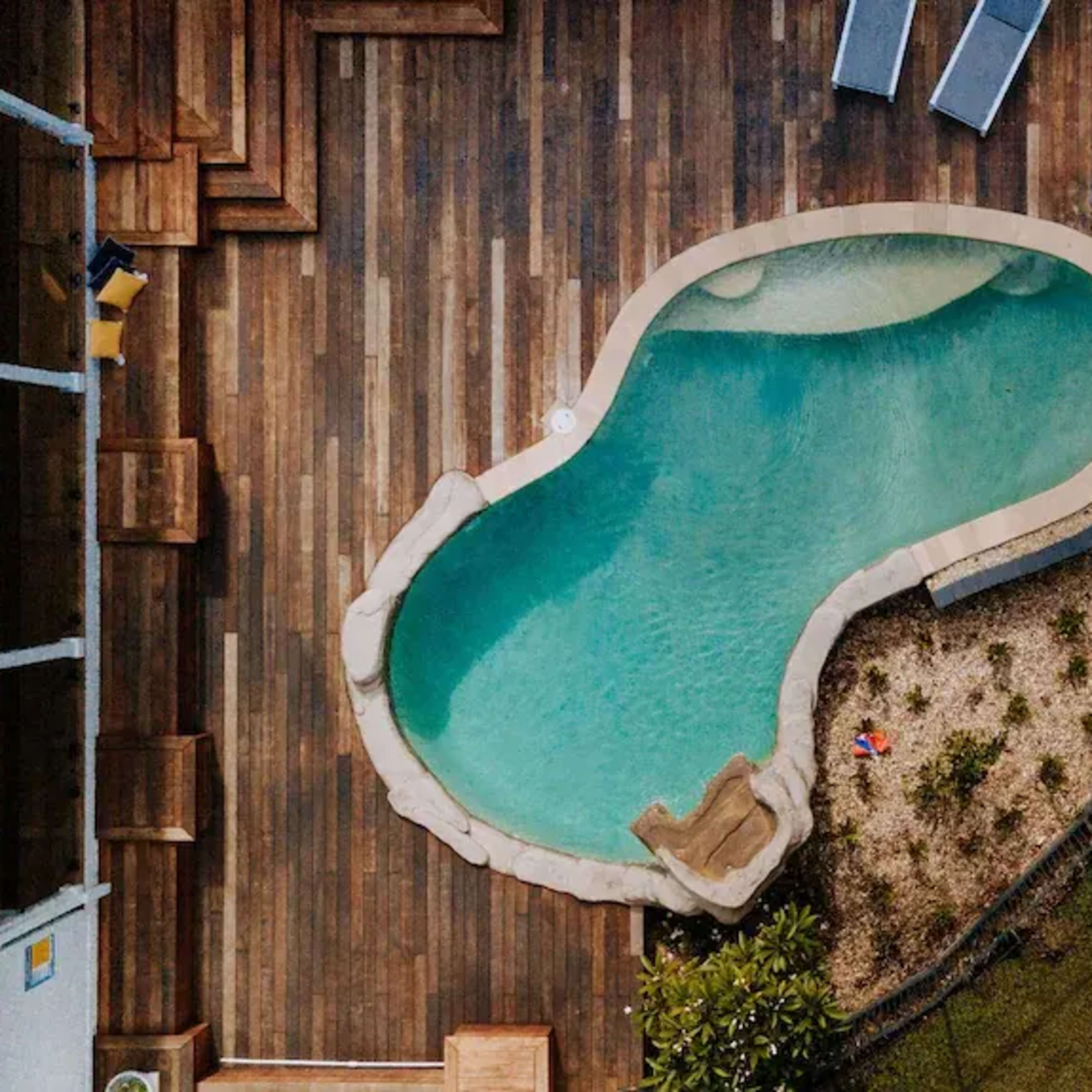 An aerial view shows a kidney-shaped swimming pool surrounded by wood decking and landscaped greenery.