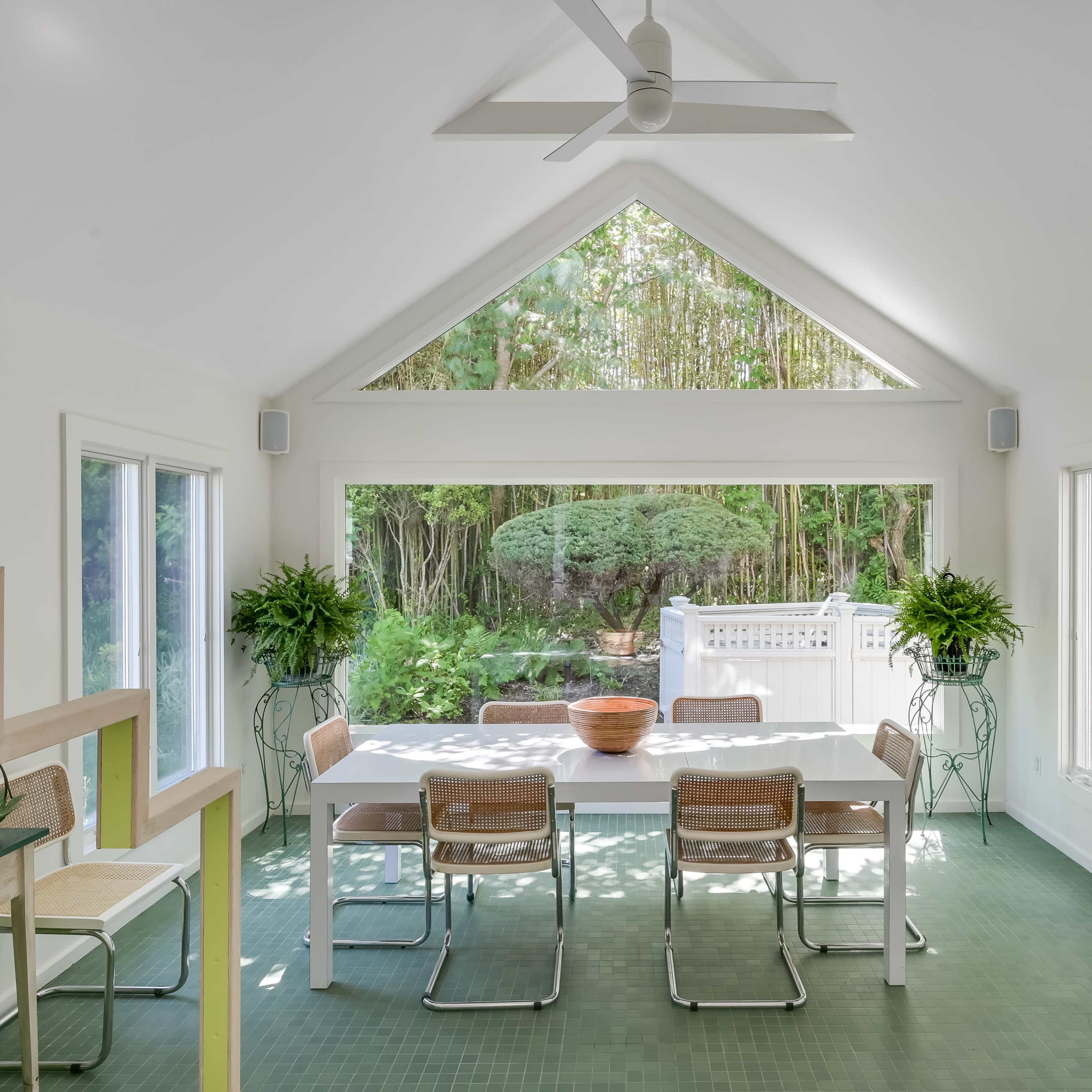 A bright dining area with a long table and chairs, surrounded by large windows that overlook a garden.
