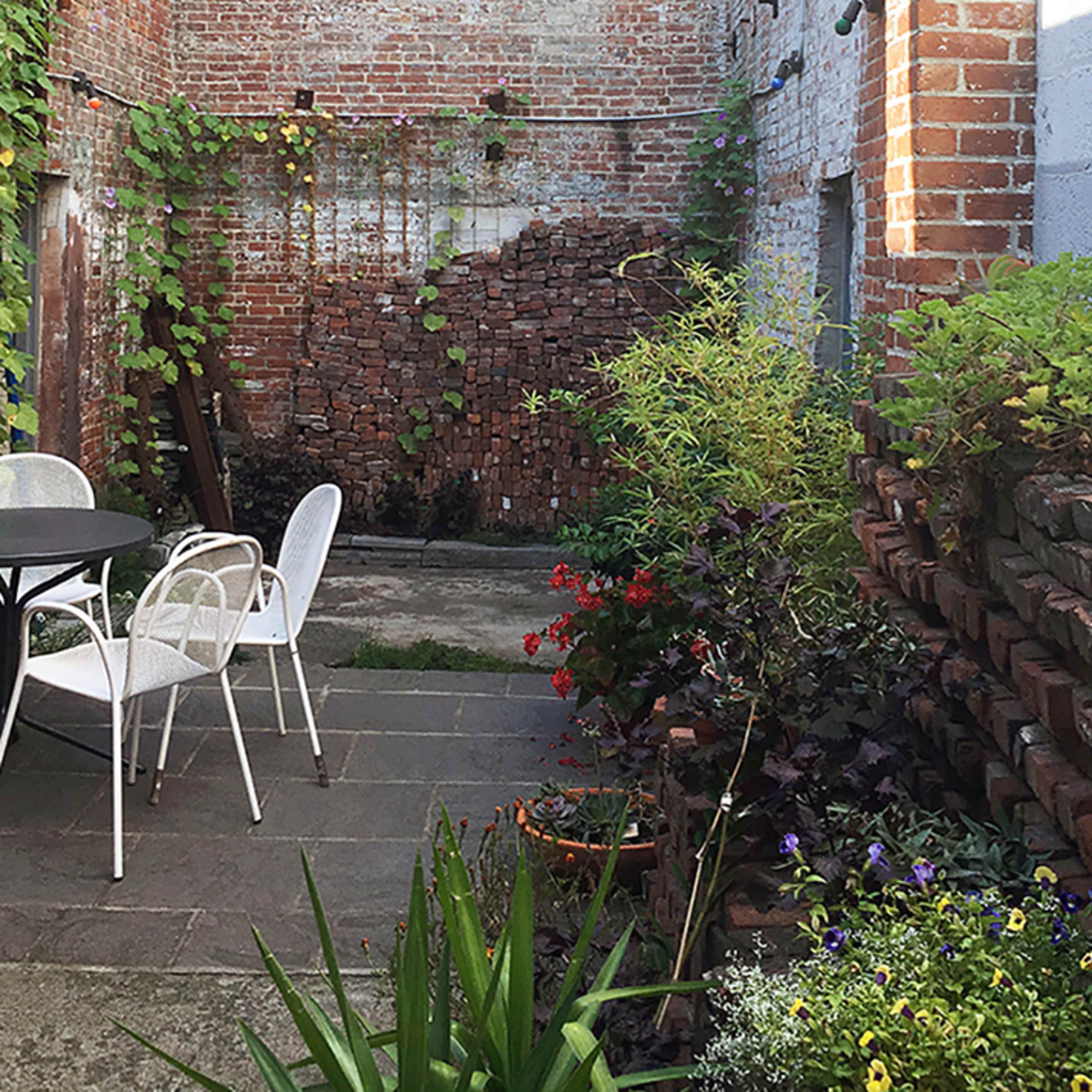A small courtyard with a round table and white chairs surrounded by brick walls and various plants along the edges.