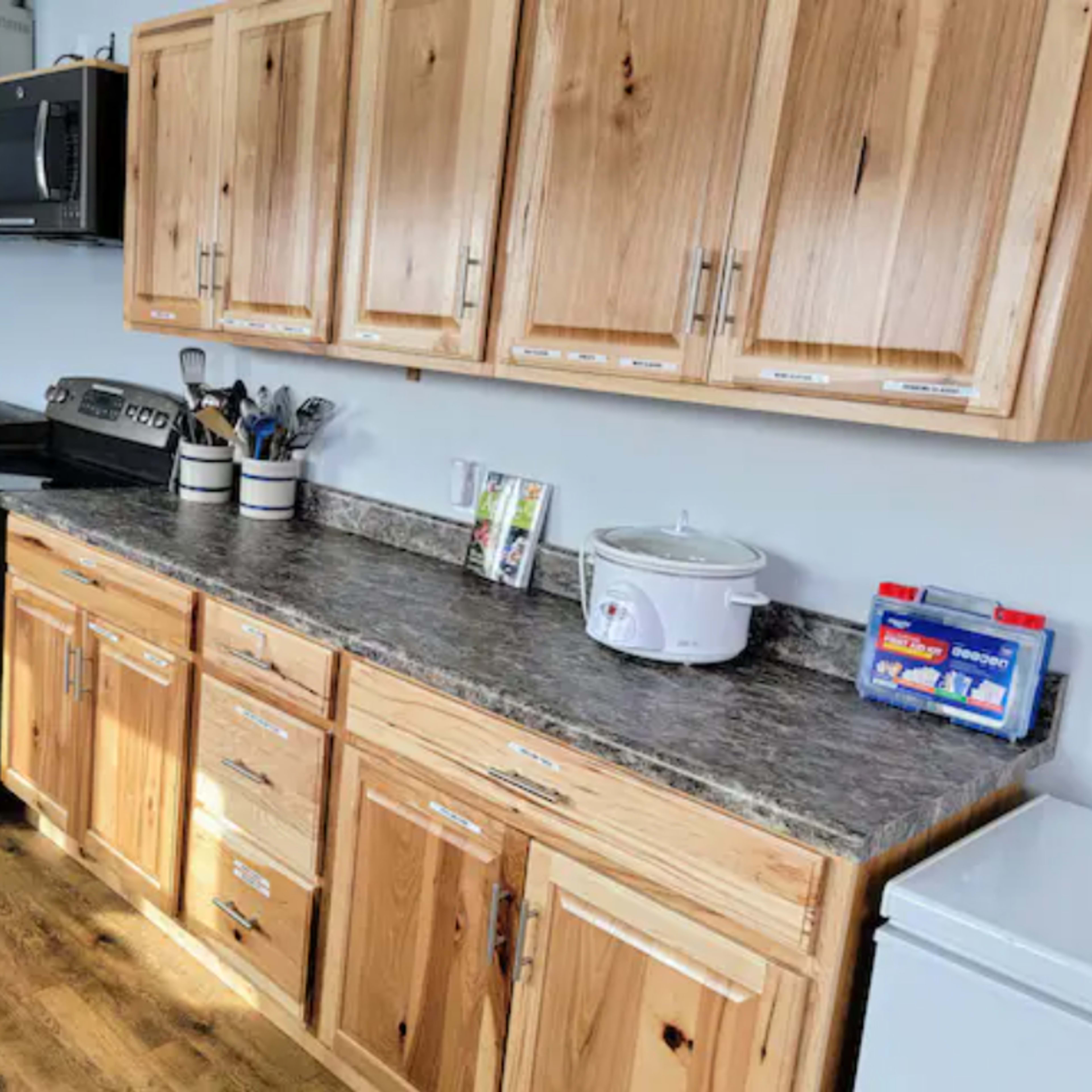 A kitchen with wooden cabinets, a stainless steel oven, a microwave above, and a countertop with cooking utensils and a slow cooker.