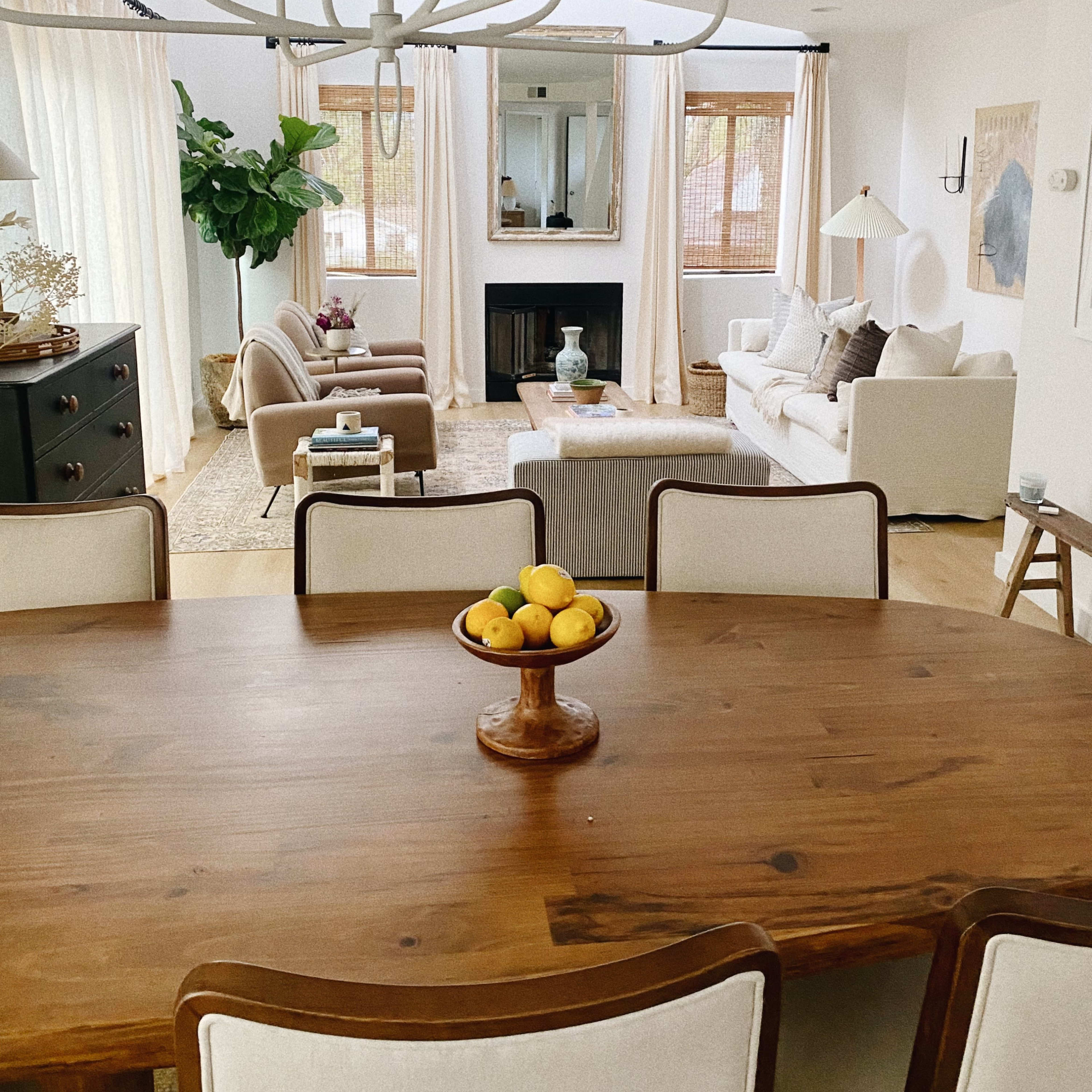 A dining table with a bowl of lemons is set in the foreground, facing a bright living room with two armchairs, a sofa, and large windows.