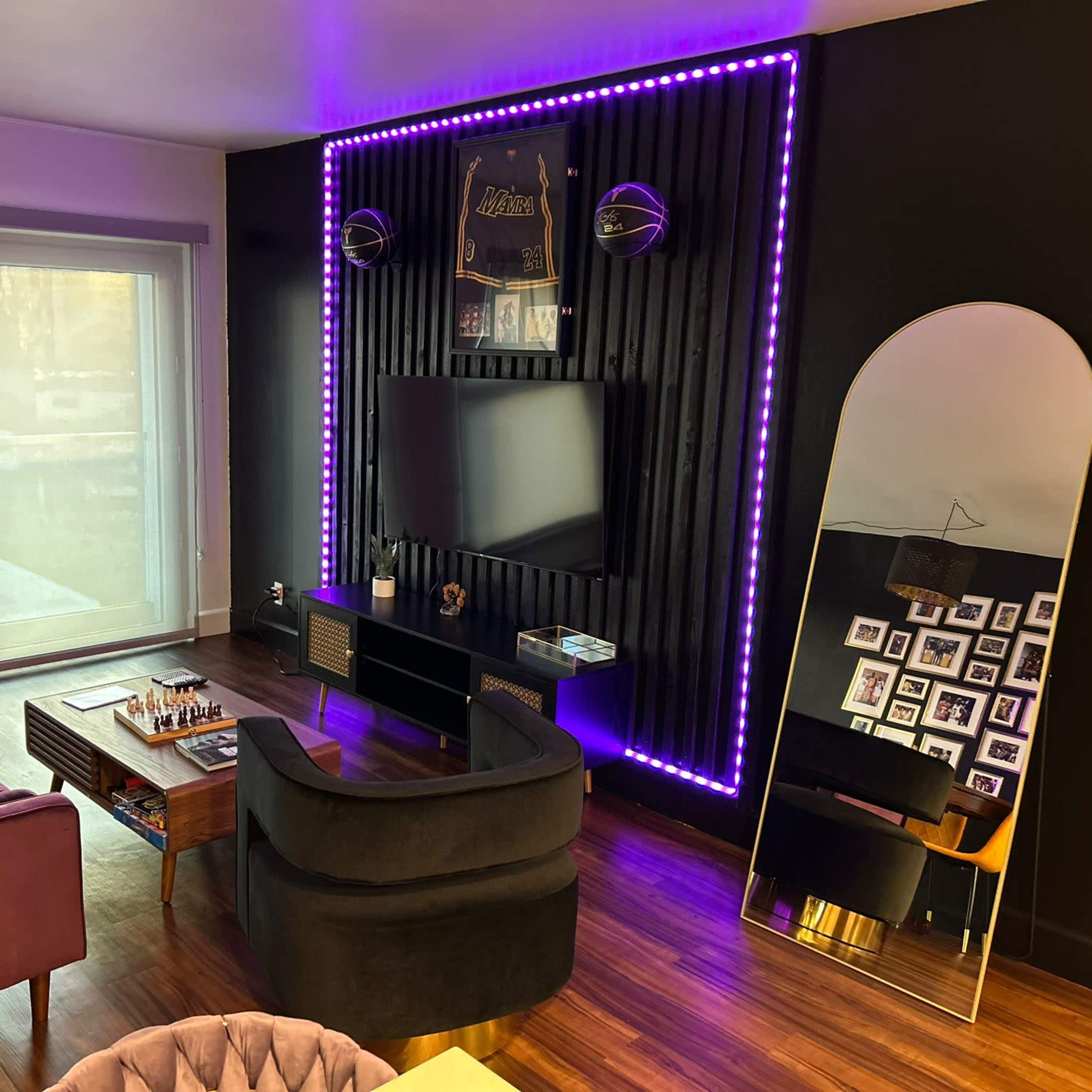 A modern living room with a purple couch, a black TV stand, an illuminated wall, and a large mirror beside a collection of framed photos.