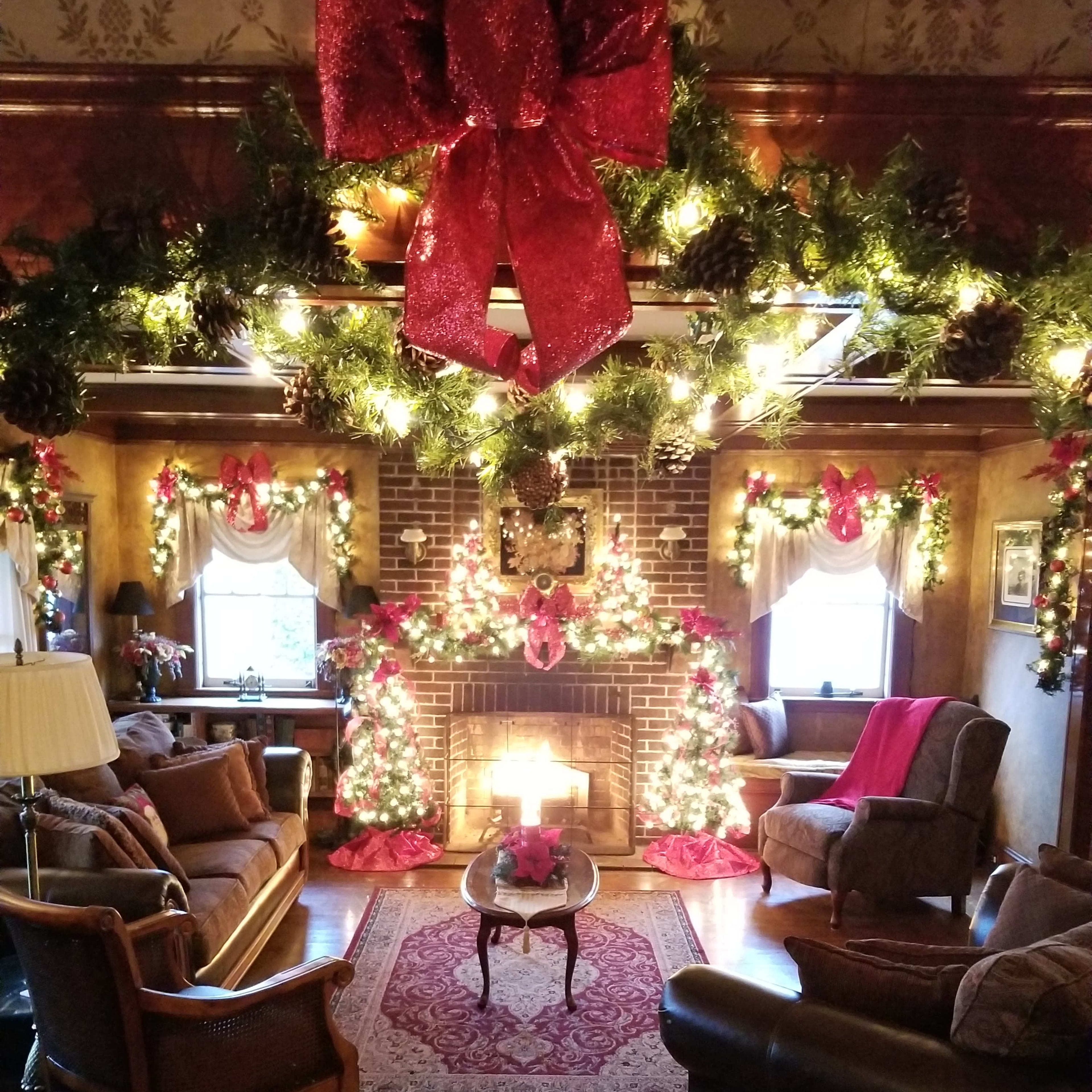 A cozy living room is decorated with festive greenery and brightly lit Christmas trees, centered around a fireplace.