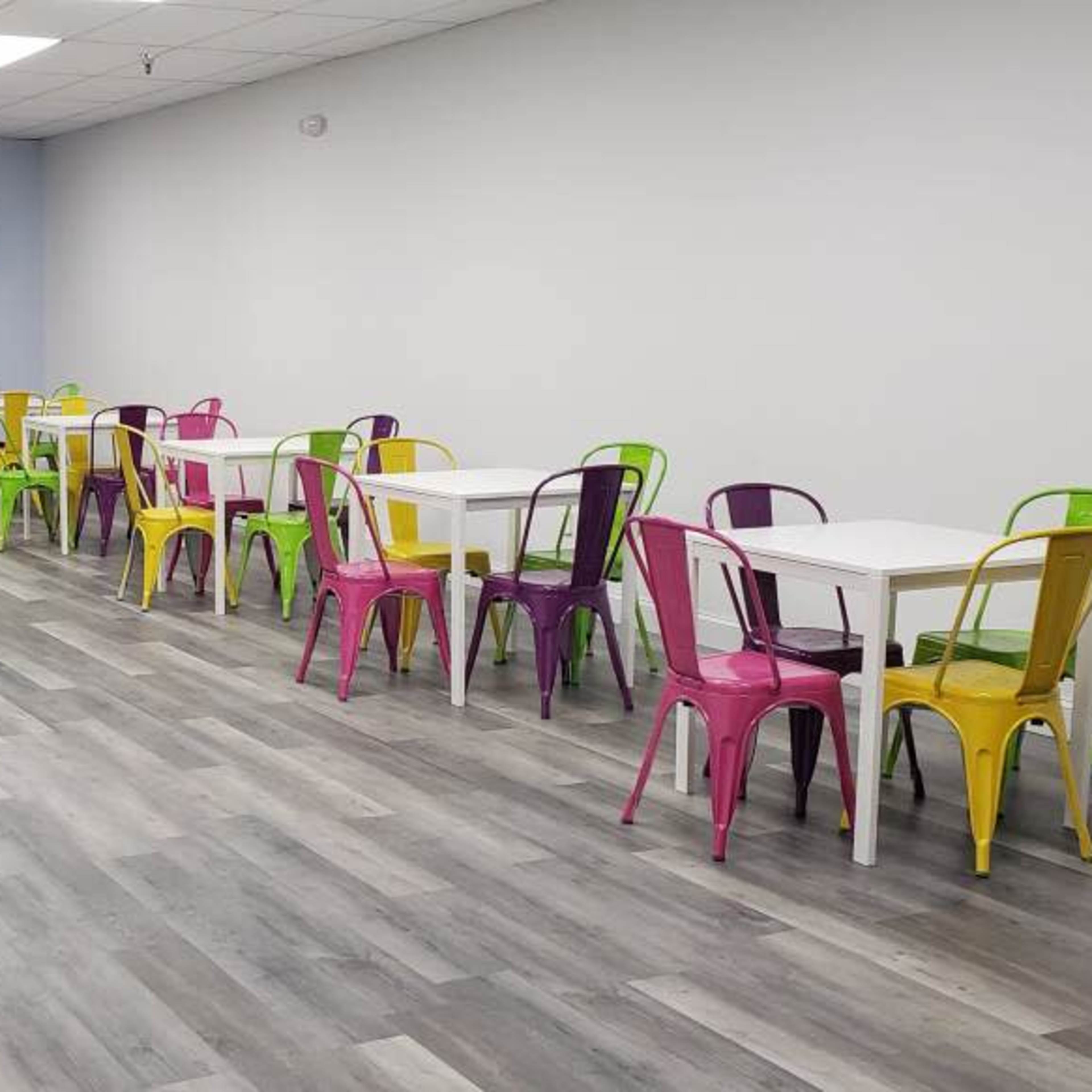 A spacious café with colorful chairs arranged around several white tables, and a counter at the front serving area.