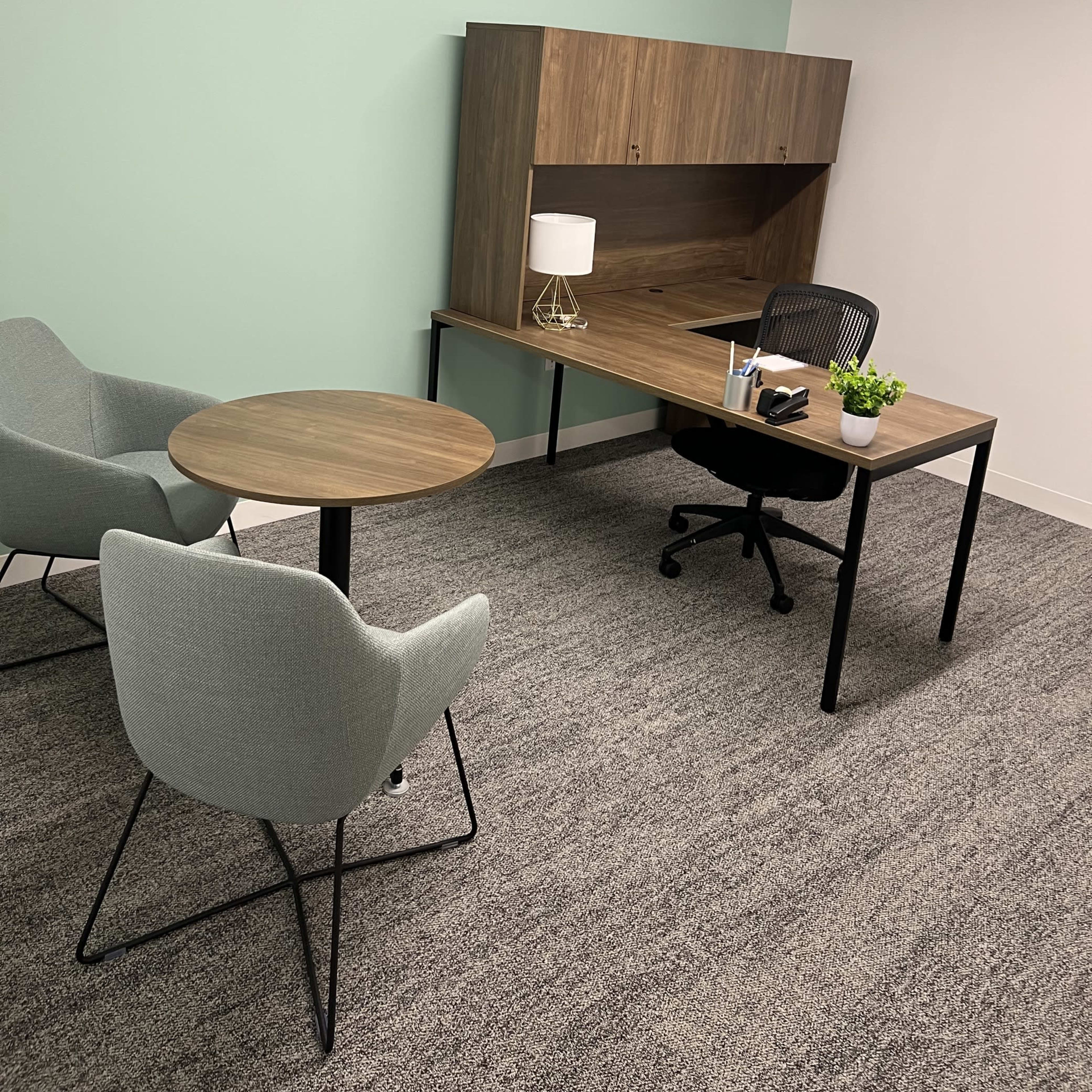 A small office space with a wooden desk, a chair, a table, and two armchairs, all set against a light green wall.