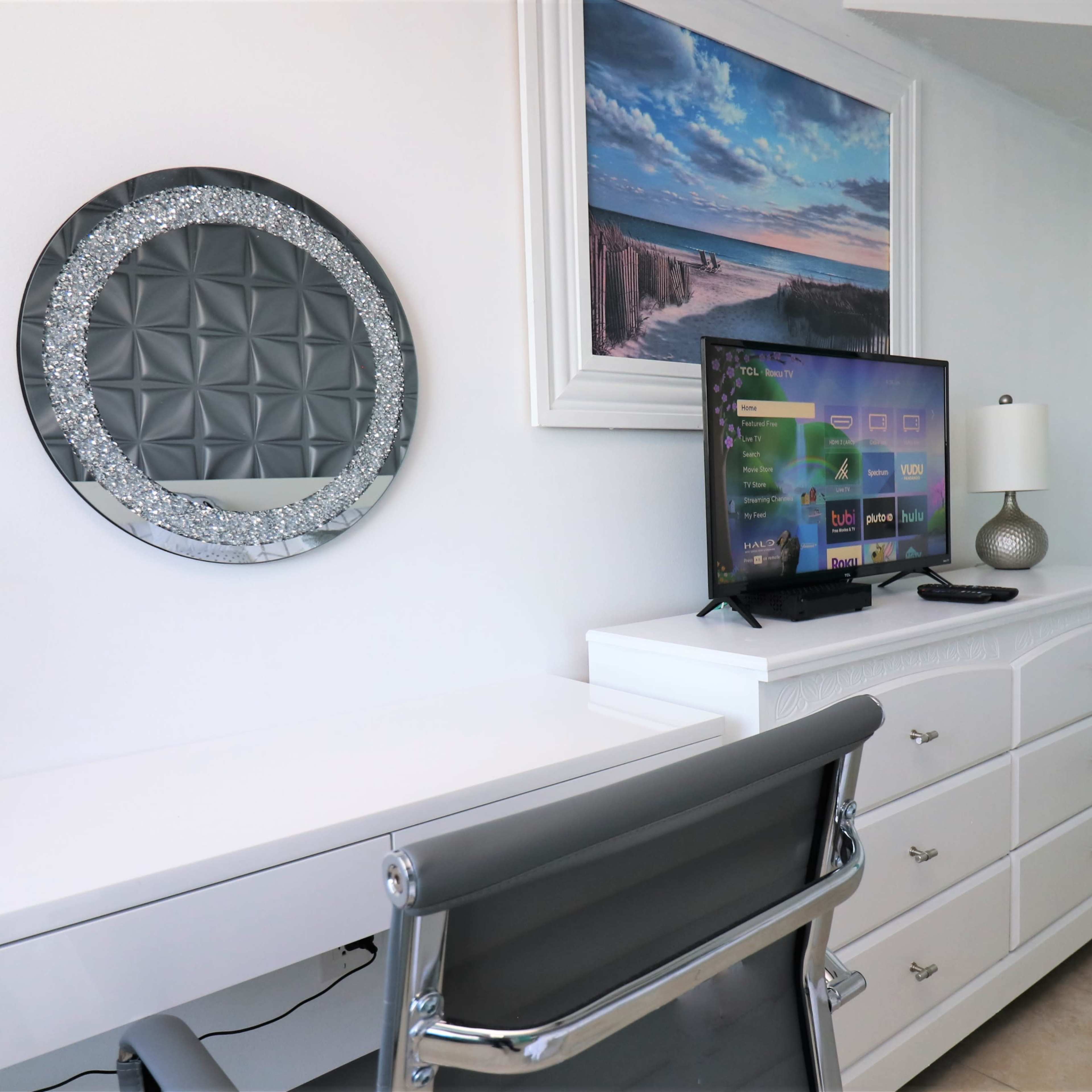 A modern, minimalist room with a white desk, a television mounted on a dresser, and a decorative mirror on the wall.