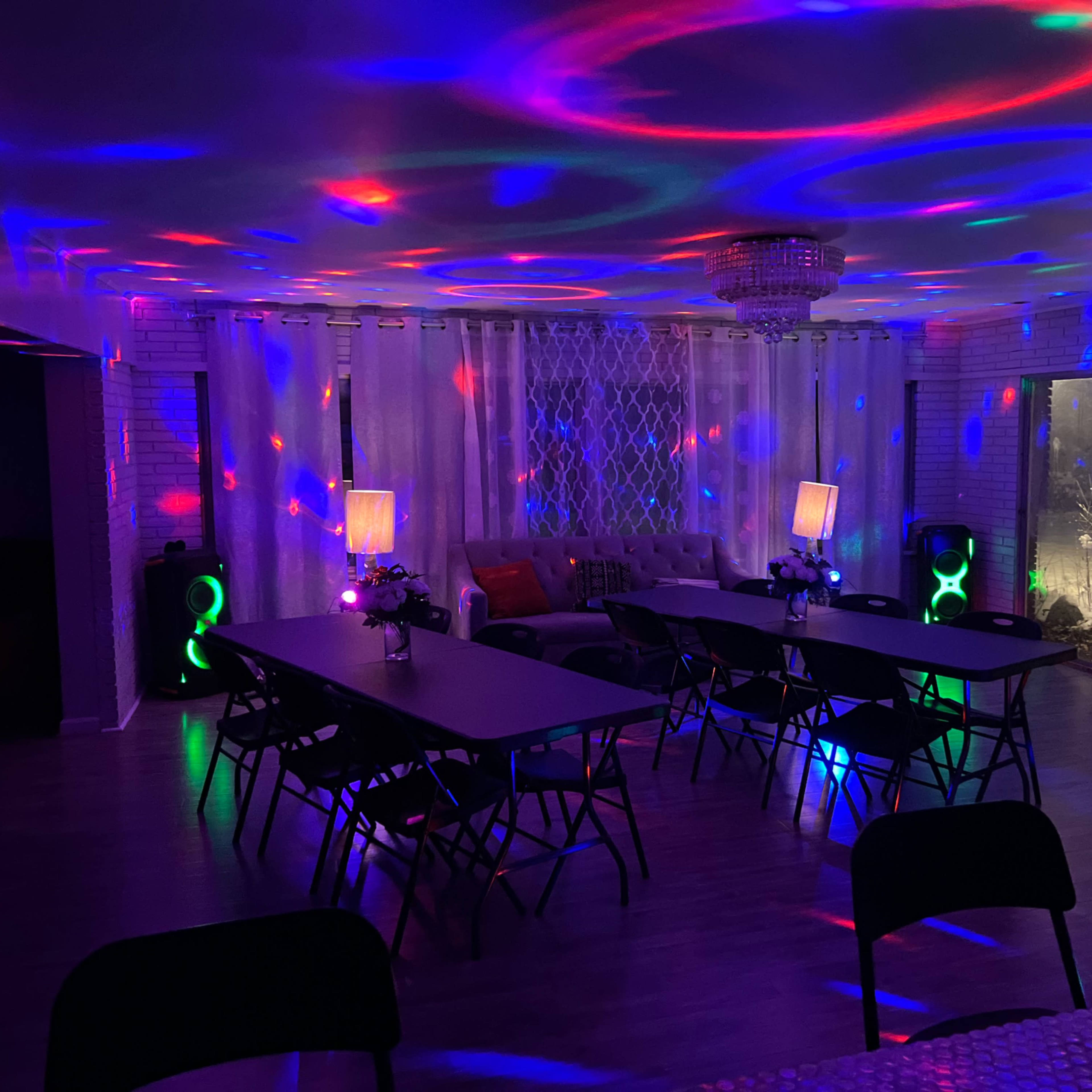 A spacious room featuring tables and chairs arranged for an event, illuminated by colorful lights and a chandelier.
