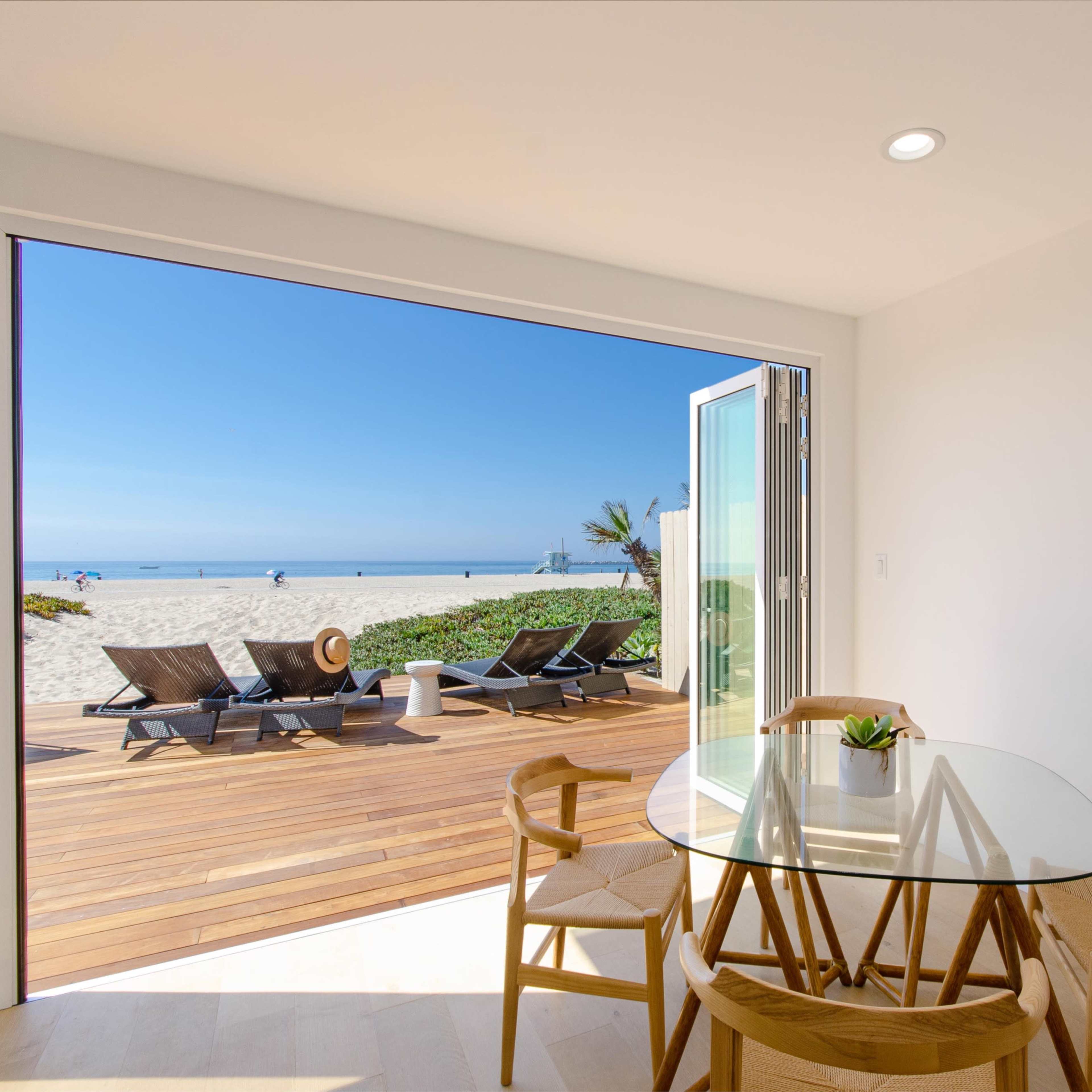 A dining area with a glass table and wooden chairs opens to a beach view through wide sliding doors.