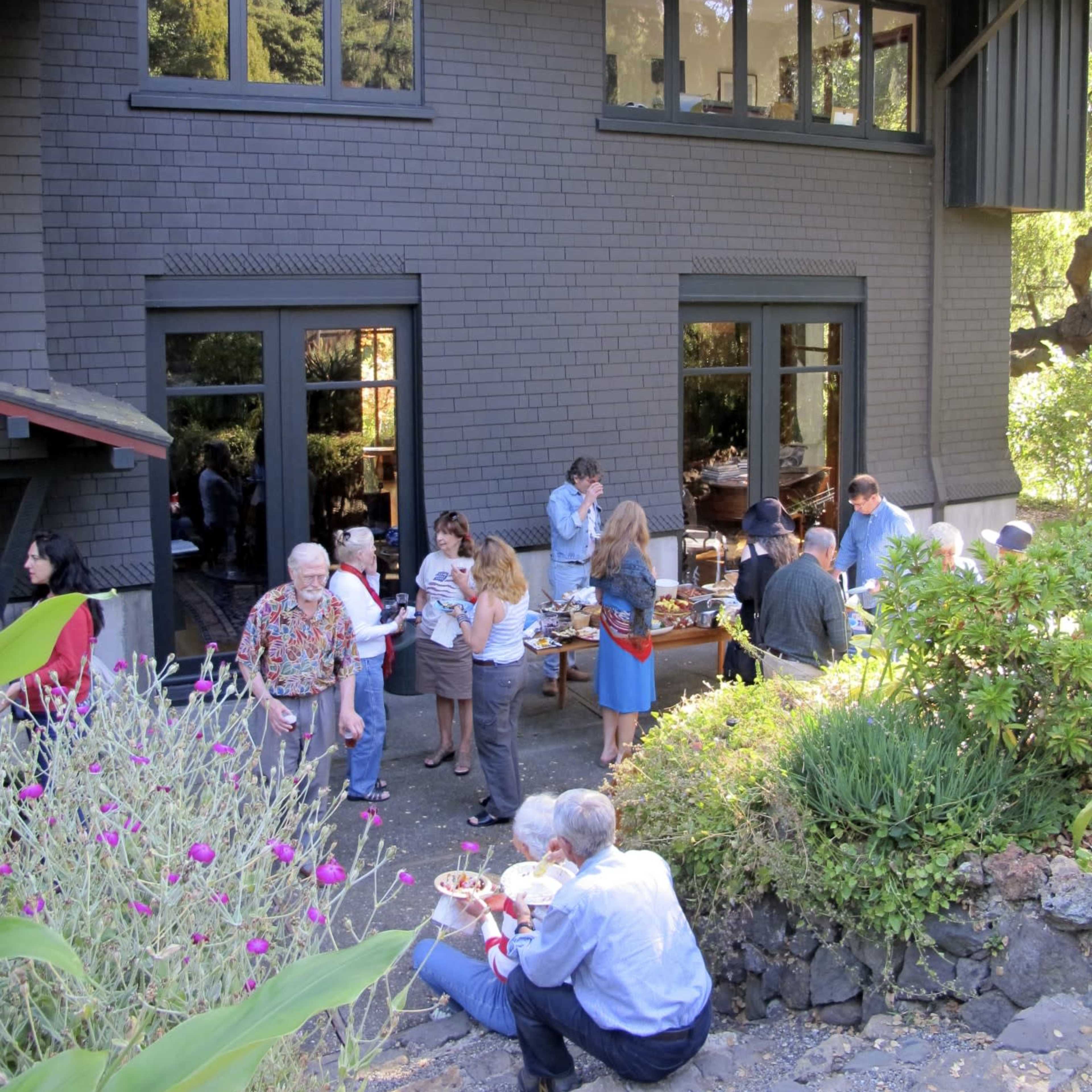 A gathering of people is taking place outdoors near a house with large windows, where attendees are enjoying food and socializing in a landscaped setting.