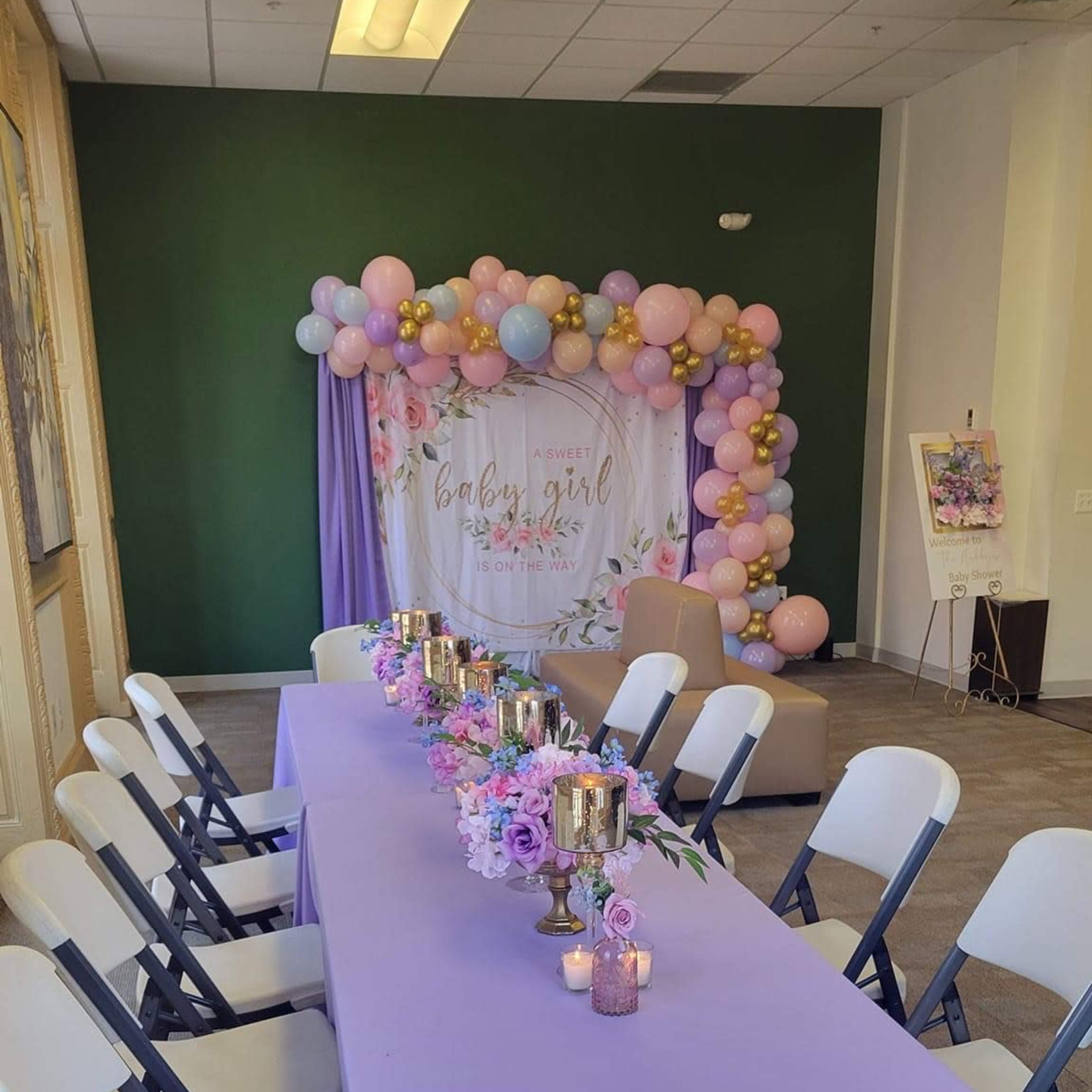 A decorated event space with a long table covered in a lavender tablecloth, floral centerpieces, and a backdrop of balloons.