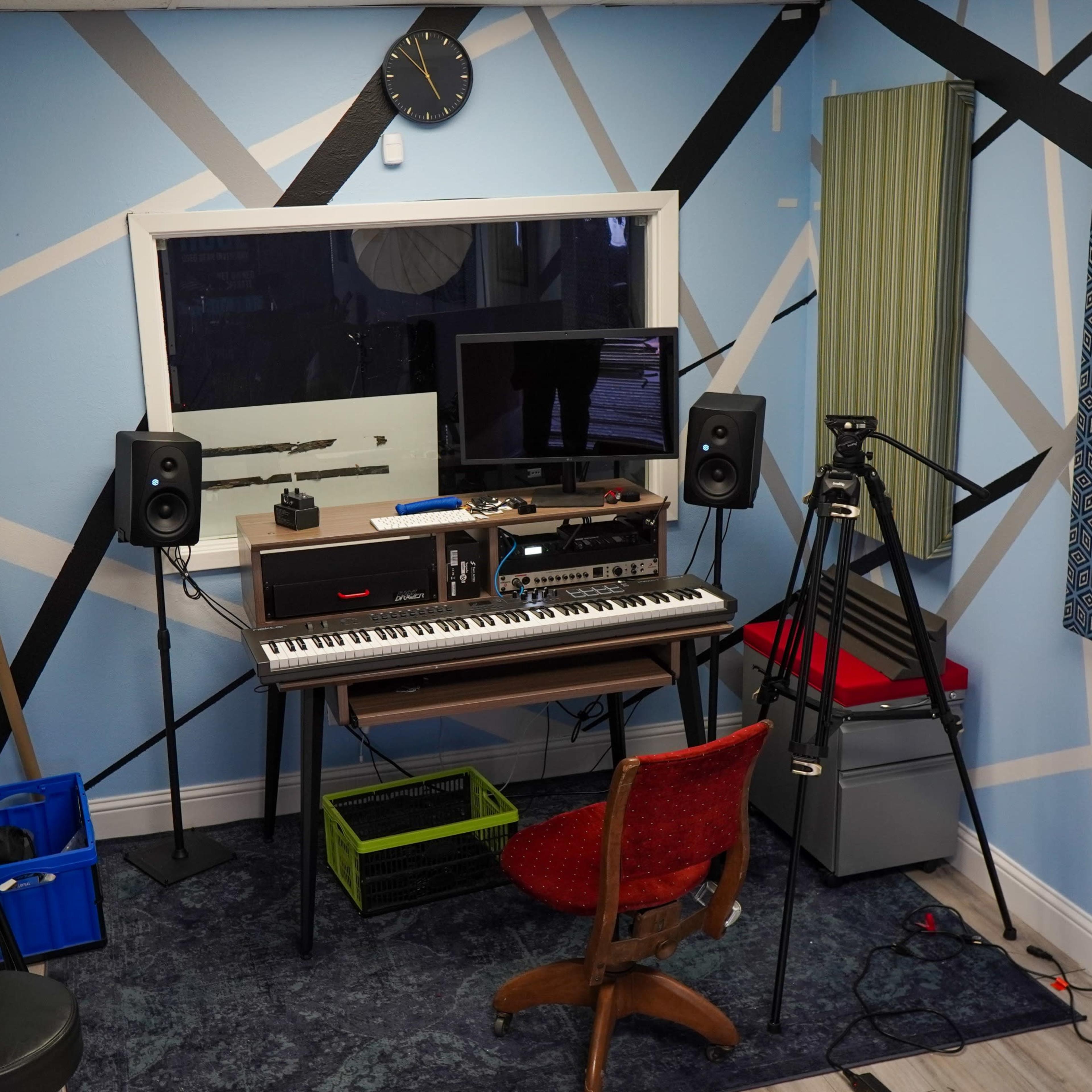 A small music studio with a keyboard, a computer setup, speakers, and various acoustic treatments on the walls, along with a chair and storage containers.