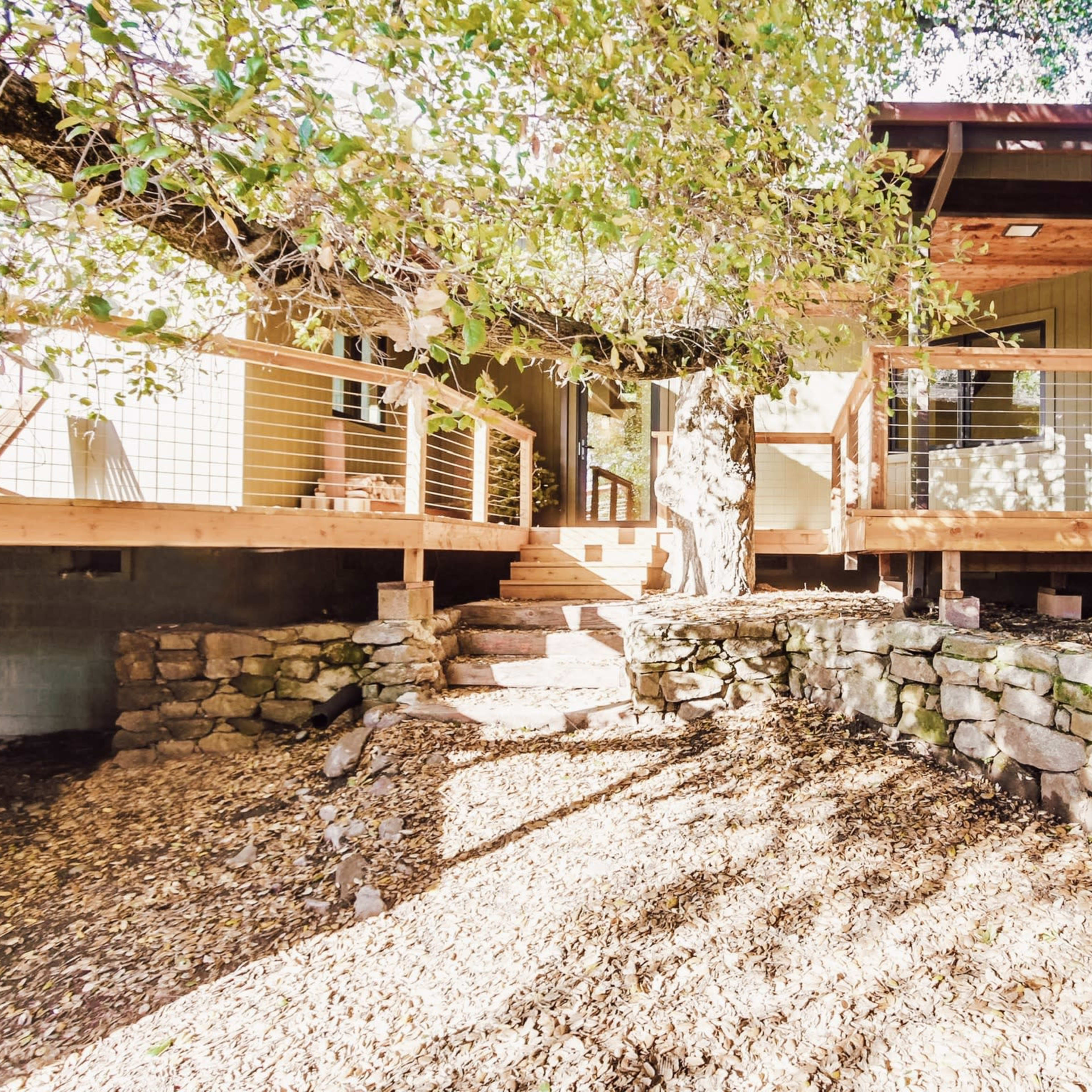 A house elevated on stone supports with wooden decks and steps leading up, surrounded by gravel and trees.
