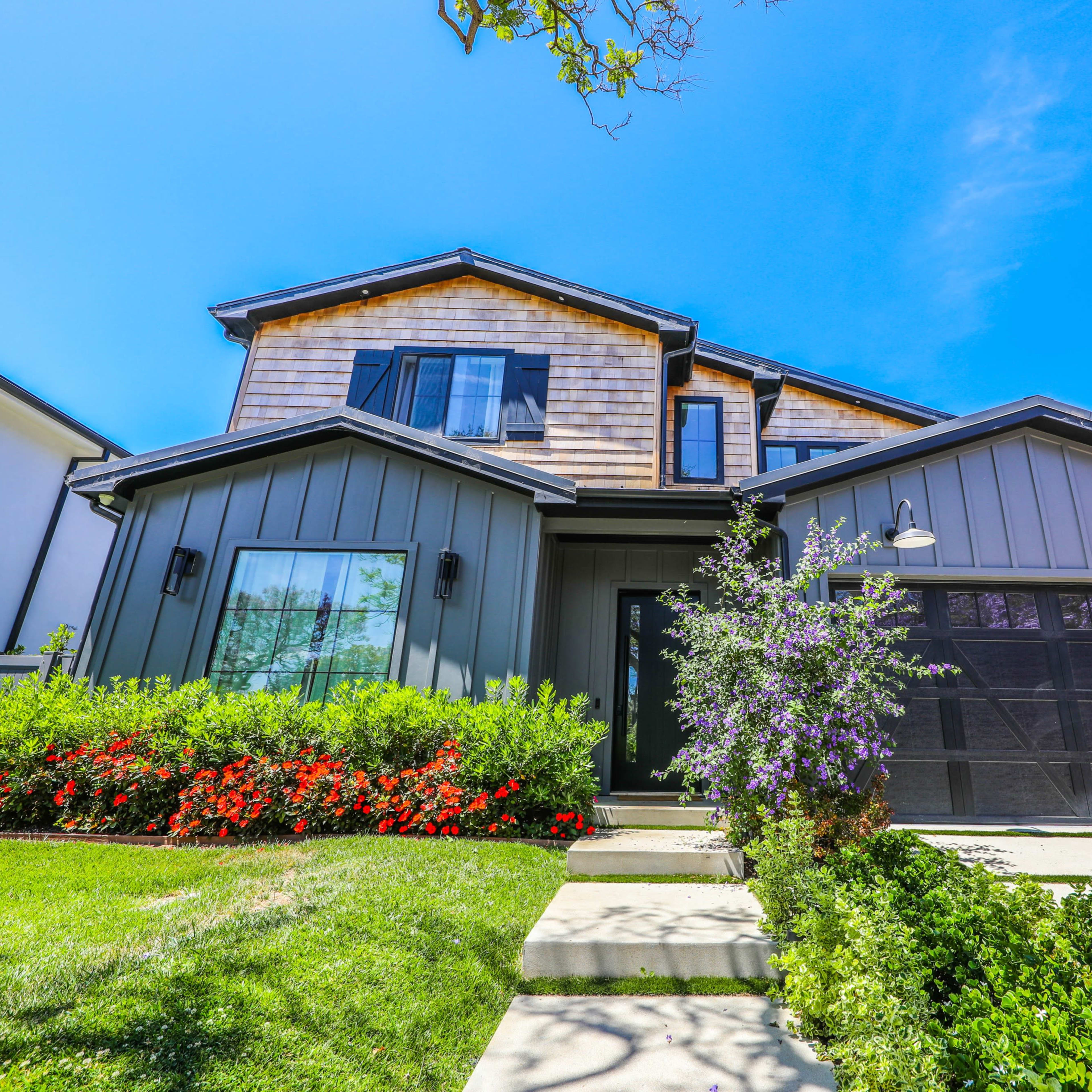 A two-story modern-style house with a gray exterior, a large front yard featuring colorful flowers, and a red car parked in the driveway.